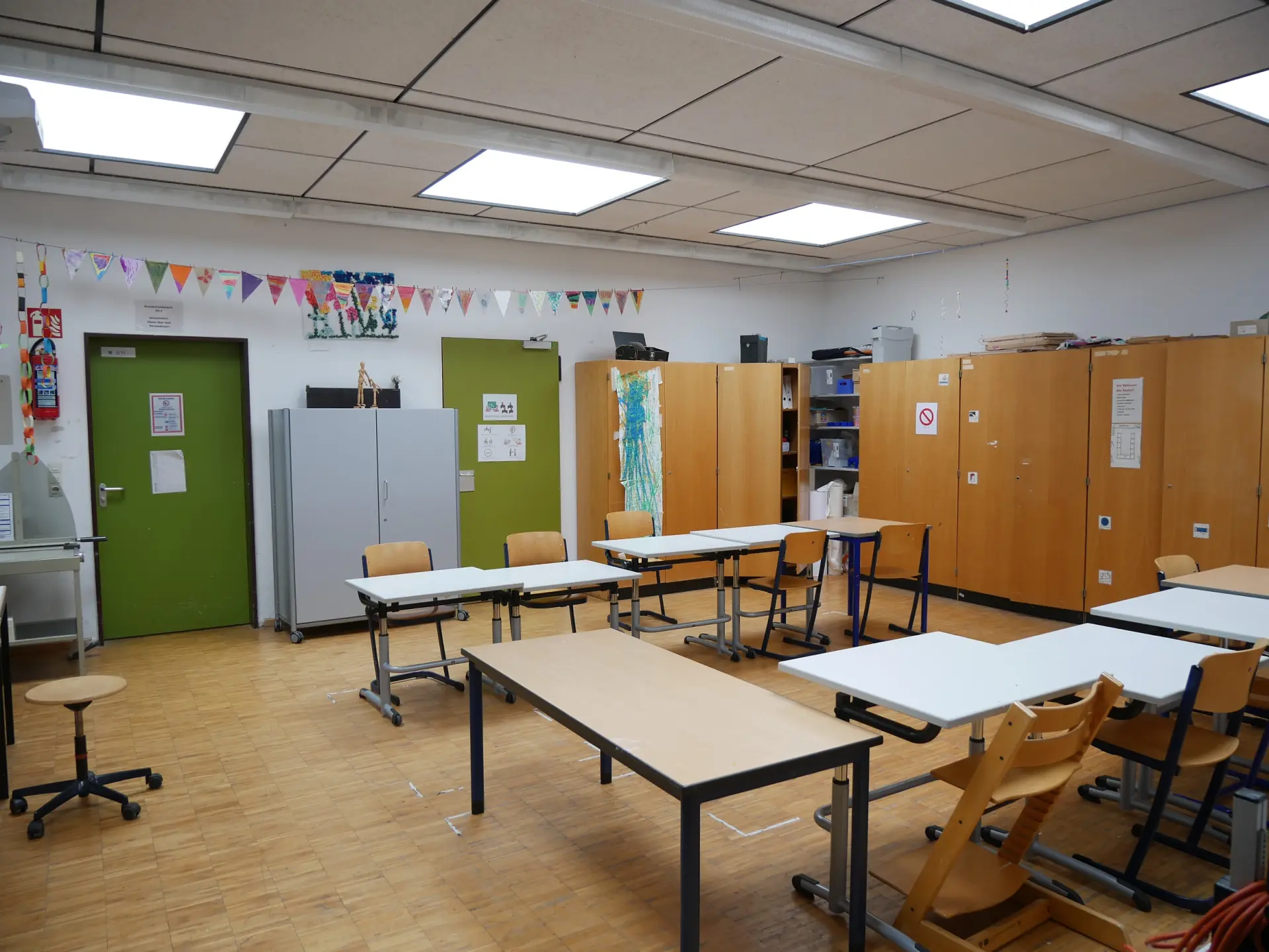 Blick in einen Klassenraum mit hellen Holzfronten, Schränken und mehreren Arbeitsstationen. Eine bunte Wimpelkette hängt an der Decke.