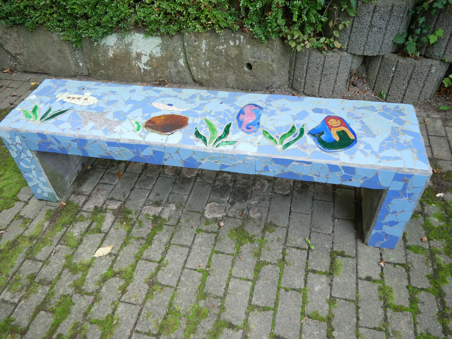 Eine mit hellblauen Mosaikfliesen verzierte Betonbank steht auf Pflastersteinen. Die Sitzfläche zeigt bunte Motive wie eine Meerjungfrau und Pflanzen.