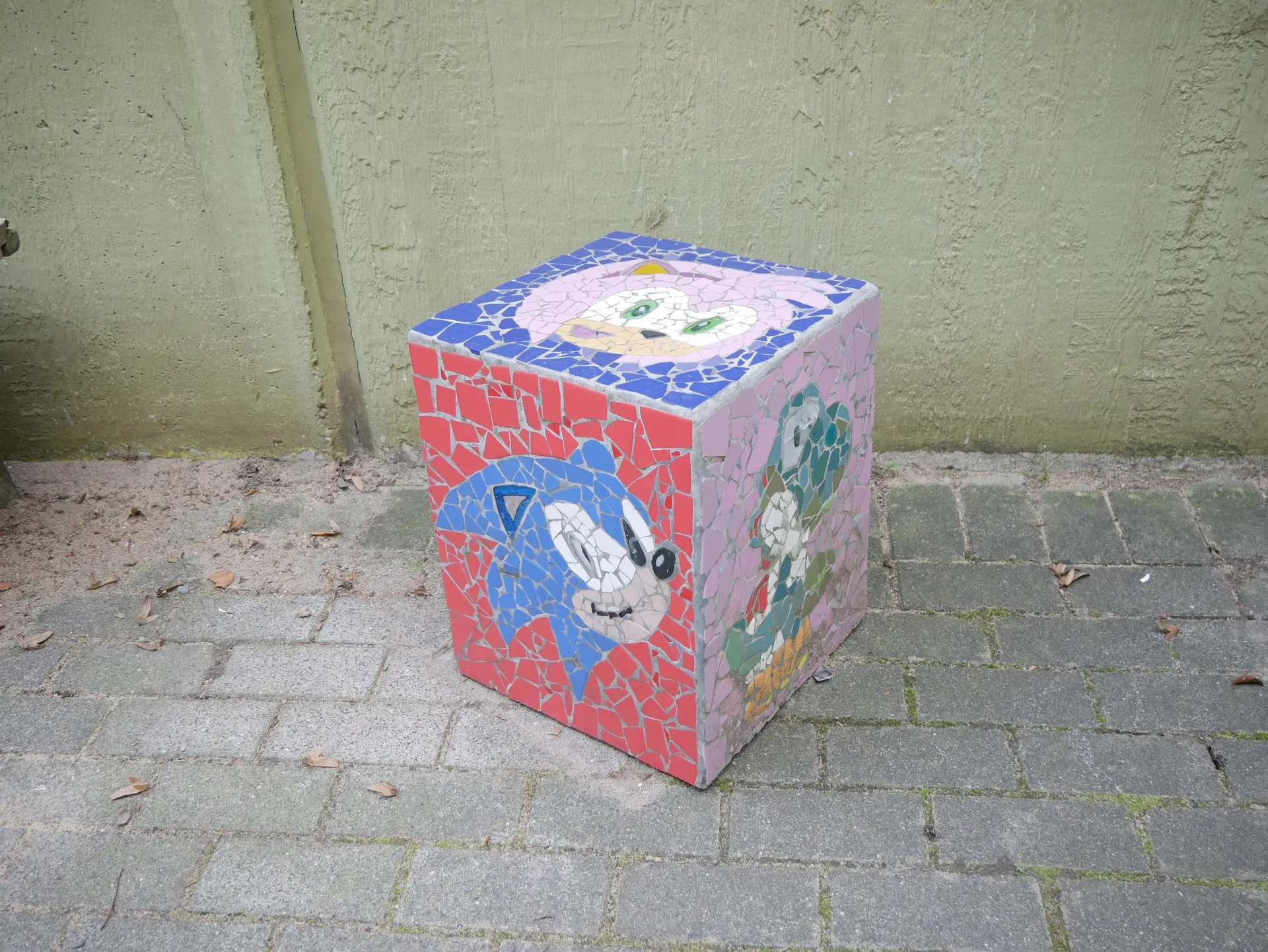 Ein Betonwürfel auf Pflastersteinen, verziert mit bunten Mosaikbildern von Sonic dem Igel und Yoshi auf rosa und roten Hintergründen.