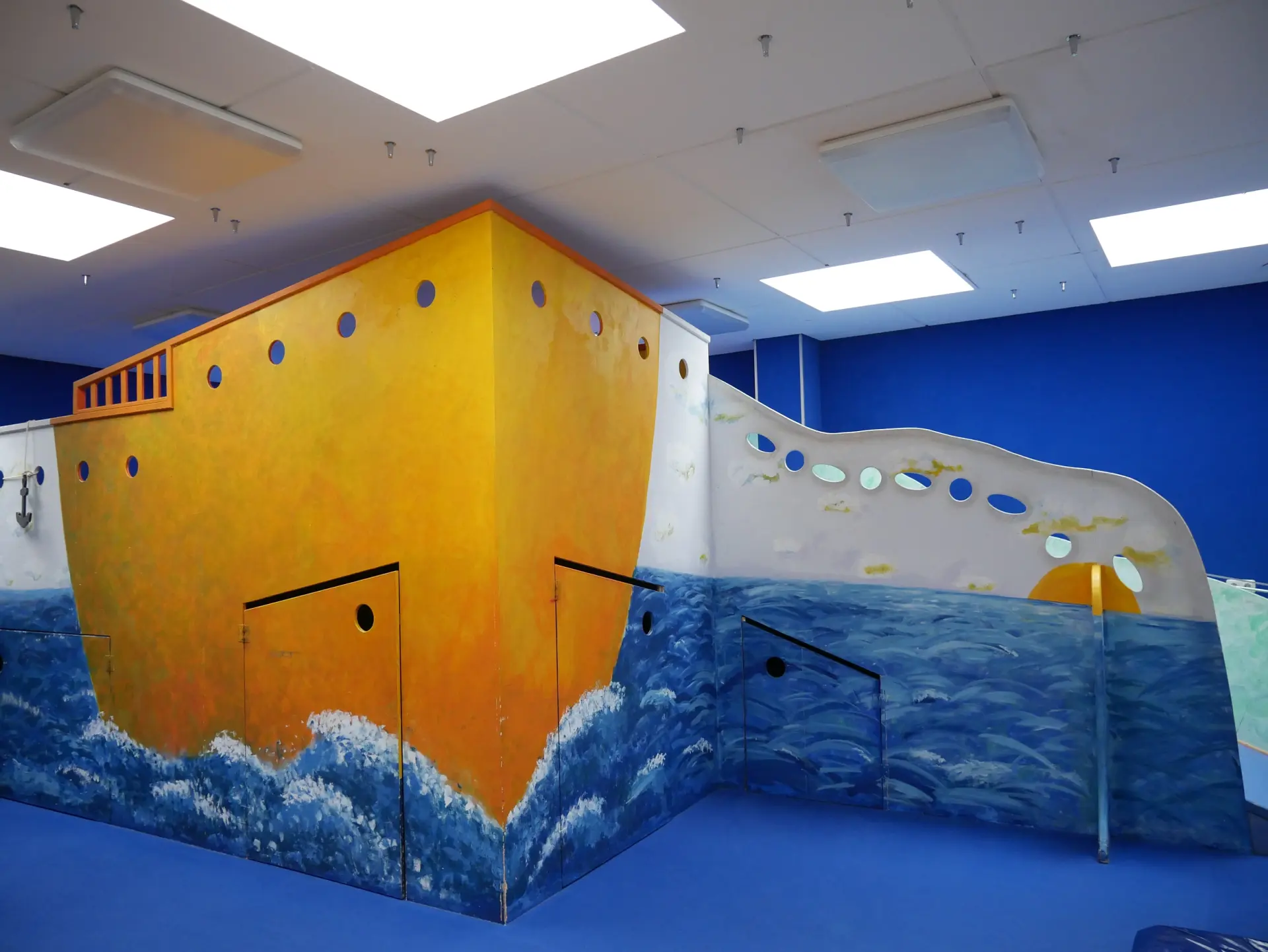 Ein großer Innenbereich mit blauem Boden, in dem ein großes, bemaltes Spielschiff steht. Die Wände sind mit blauen Wellen und einem orange-weißen Schiffsrumpf bemalt.