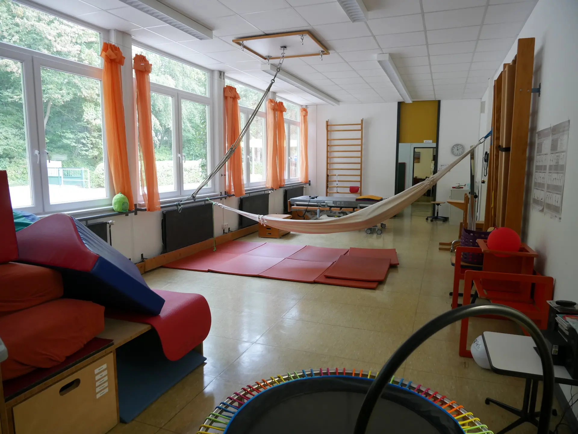 Ein Behandlungsraum mit großen Fenstern, orangefarbenen Vorhängen, einer Hängematte, Sprossenwand, Matten und Turngeräten.