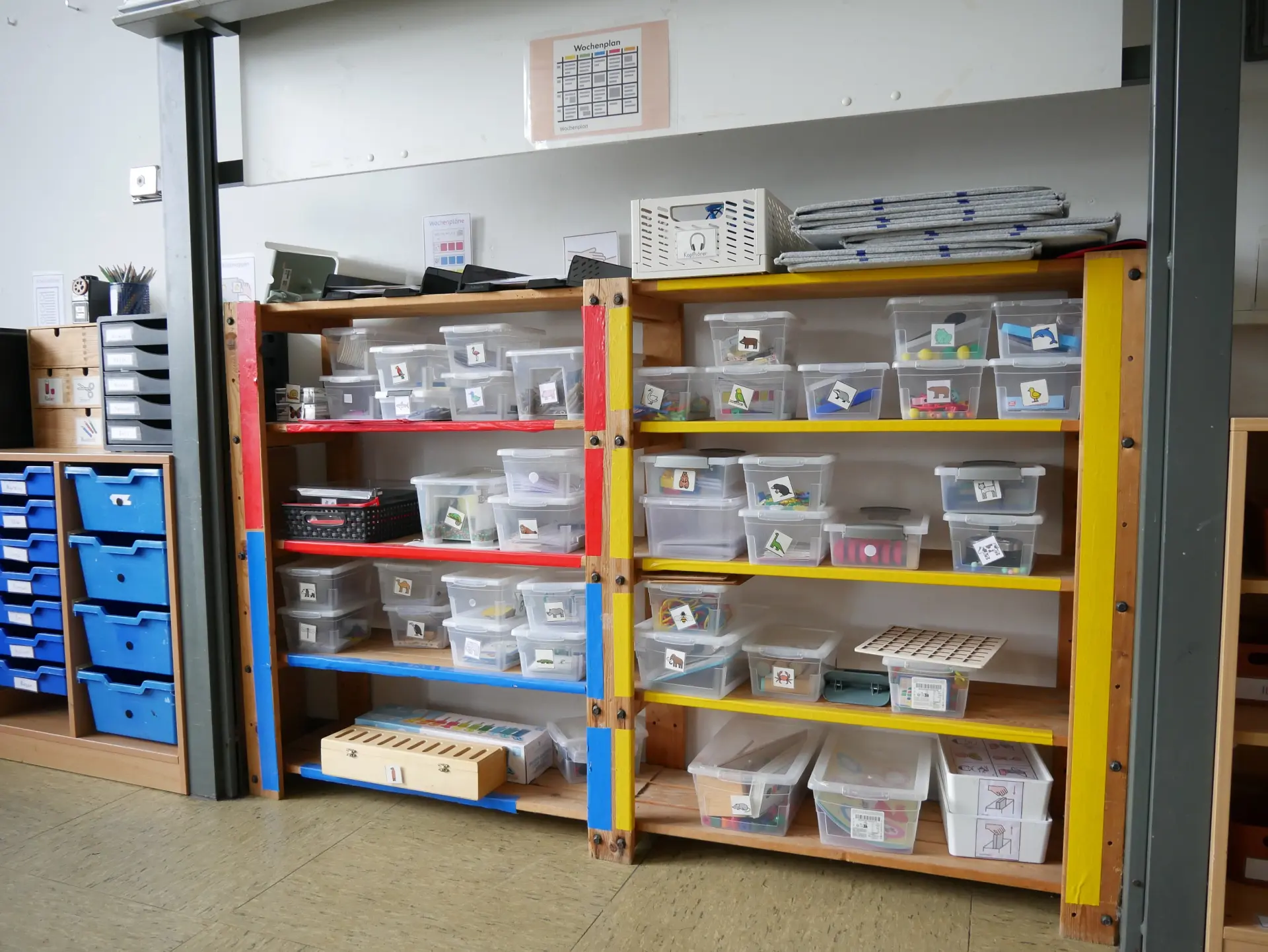 Buntes Holzregal im Klassenzimmer, gefüllt mit vielen beschrifteten, transparenten Kunststoffboxen für Lernmaterialien.