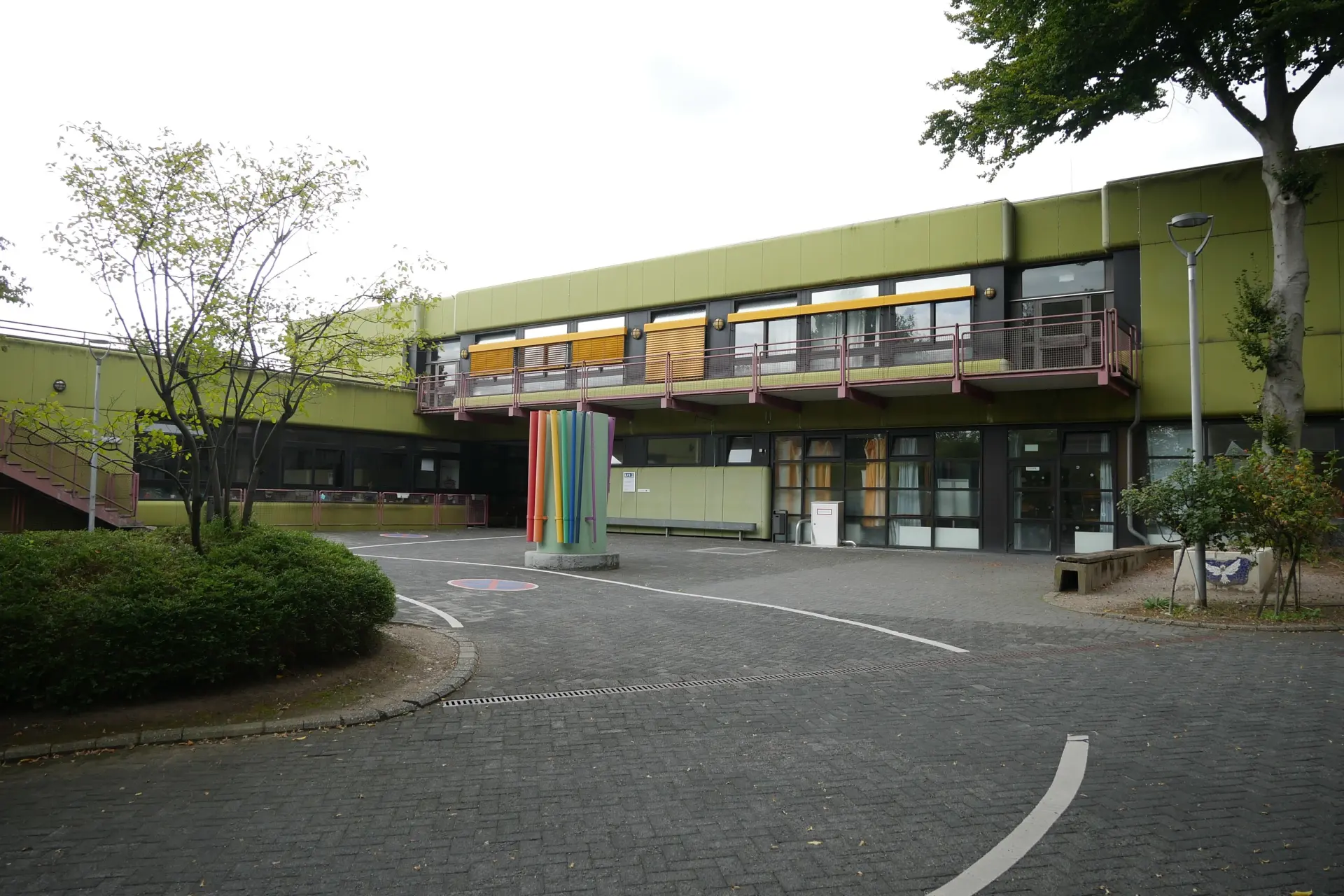 Blick auf das Schulgebäude: ein Flachbau in grüner Betonoptik. Im Vordergrund ein Schulhof mit einem Pflanzbeet.