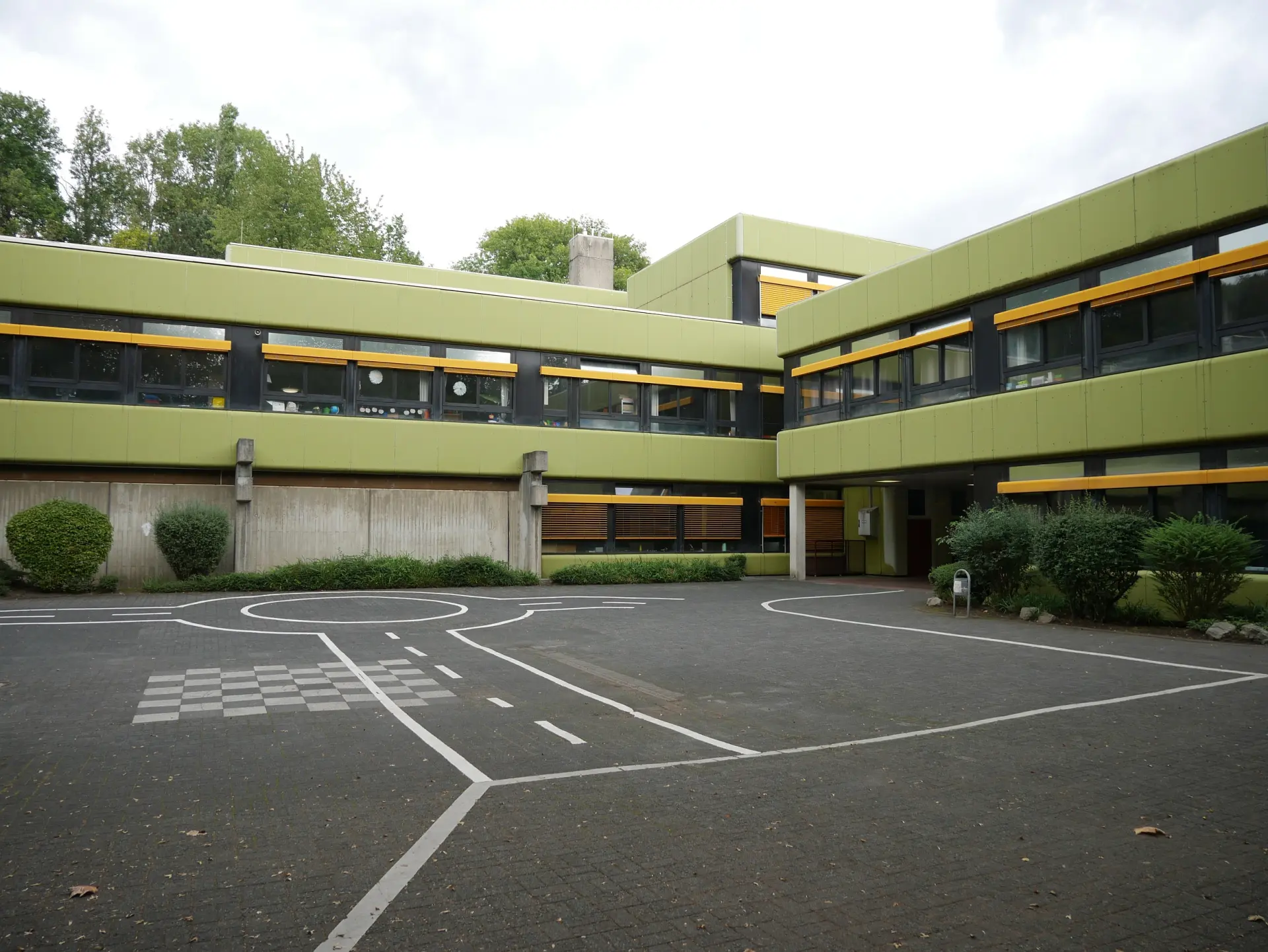 Blick auf das zweigeschossige, grüne Schulgebäude. Auf dem davor liegenden gepflasterten Hof sind Straßenmarkierungen aufgemalt.