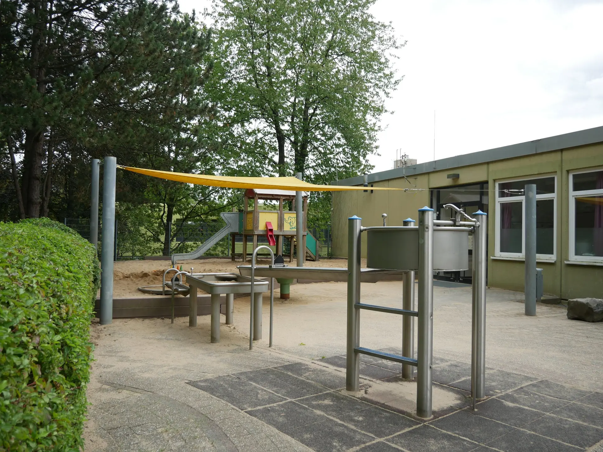 Schulhofbereich mit Edelstahl-Matsch- und Wasserspielgeräten, Sandfläche, Klettergerüst und gelbem Sonnensegel.