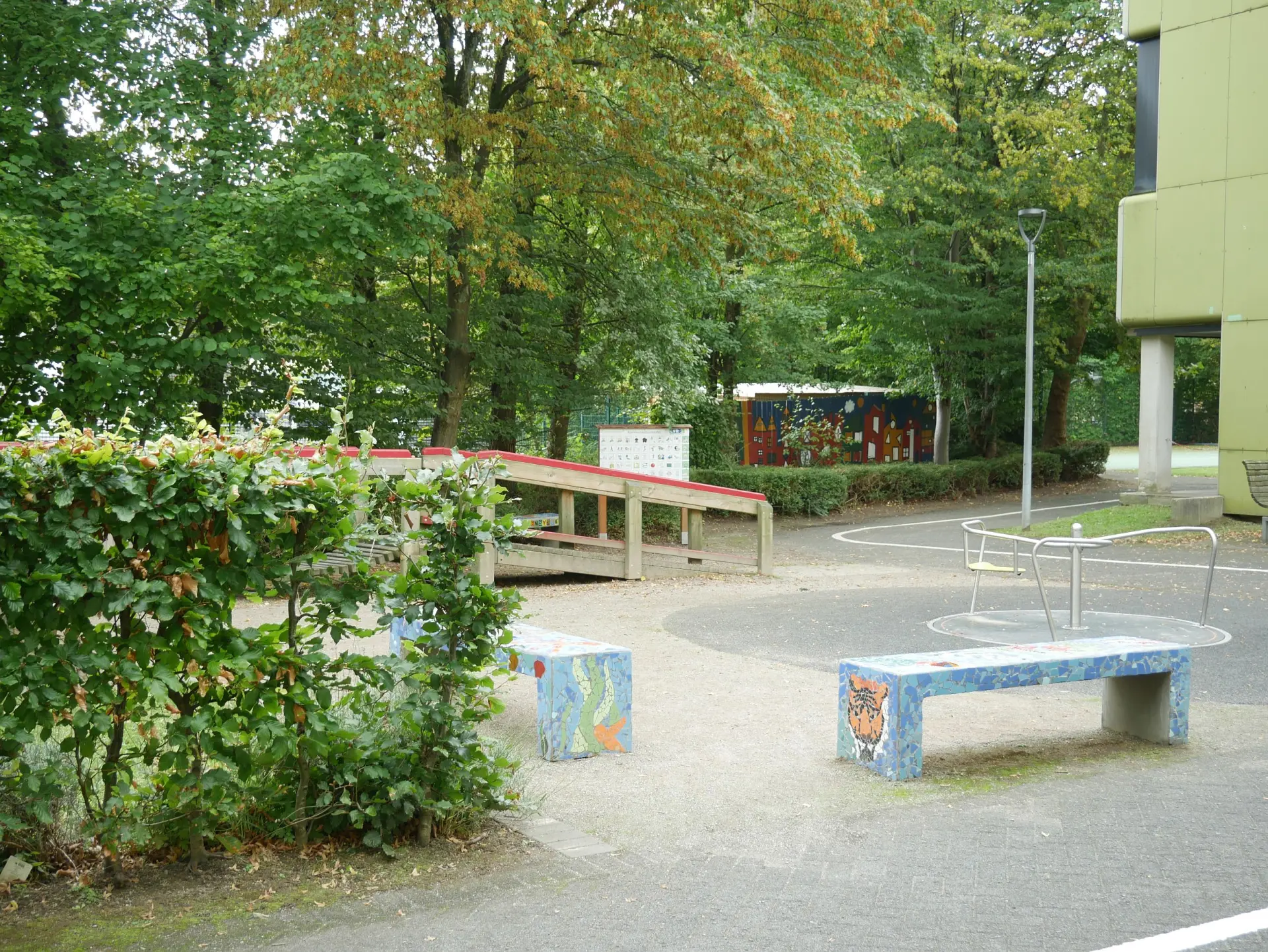 Grüner Außenbereich mit Bäumen, zwei bunt gefliesten Mosaikbänken, einer Holzrampe und einem kleinen Karussell.