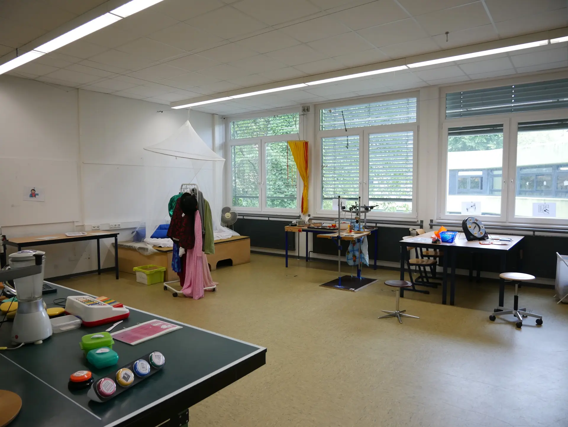 Heller Schulraum mit großen Fenstern, verschiedenen Tischen, Hilfsmitteln, Taster-Schaltern auf einem Tisch und einer Verkleidungsecke.