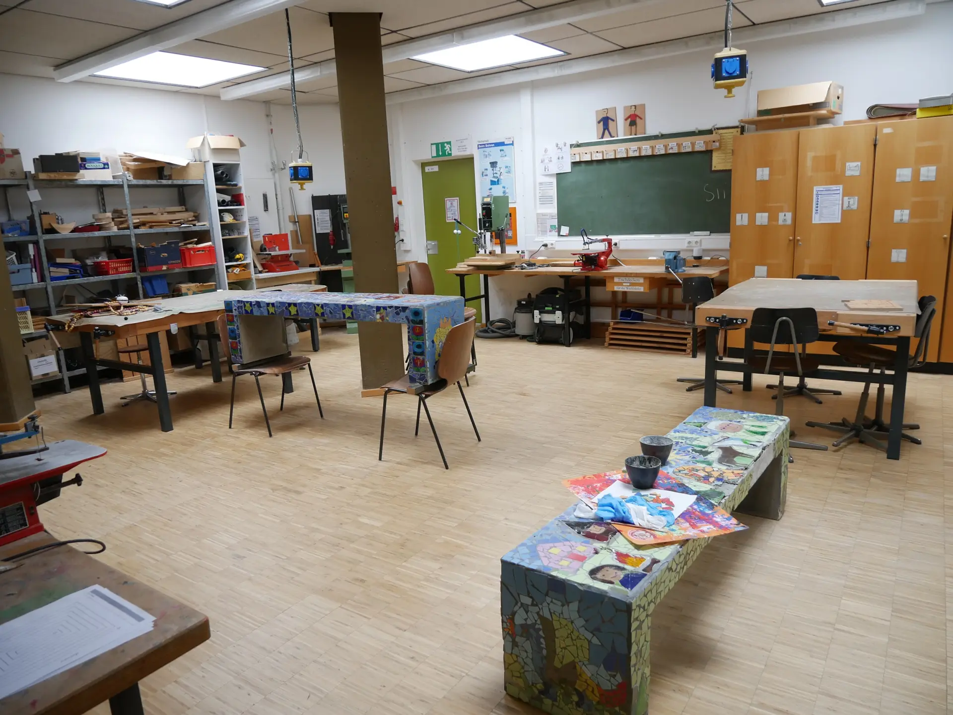 Geräumiger Werkraum mit Werktischen, Maschinen, Materialregalen, Tafel und einer bunt verzierten Mosaikbank im Vordergrund.
