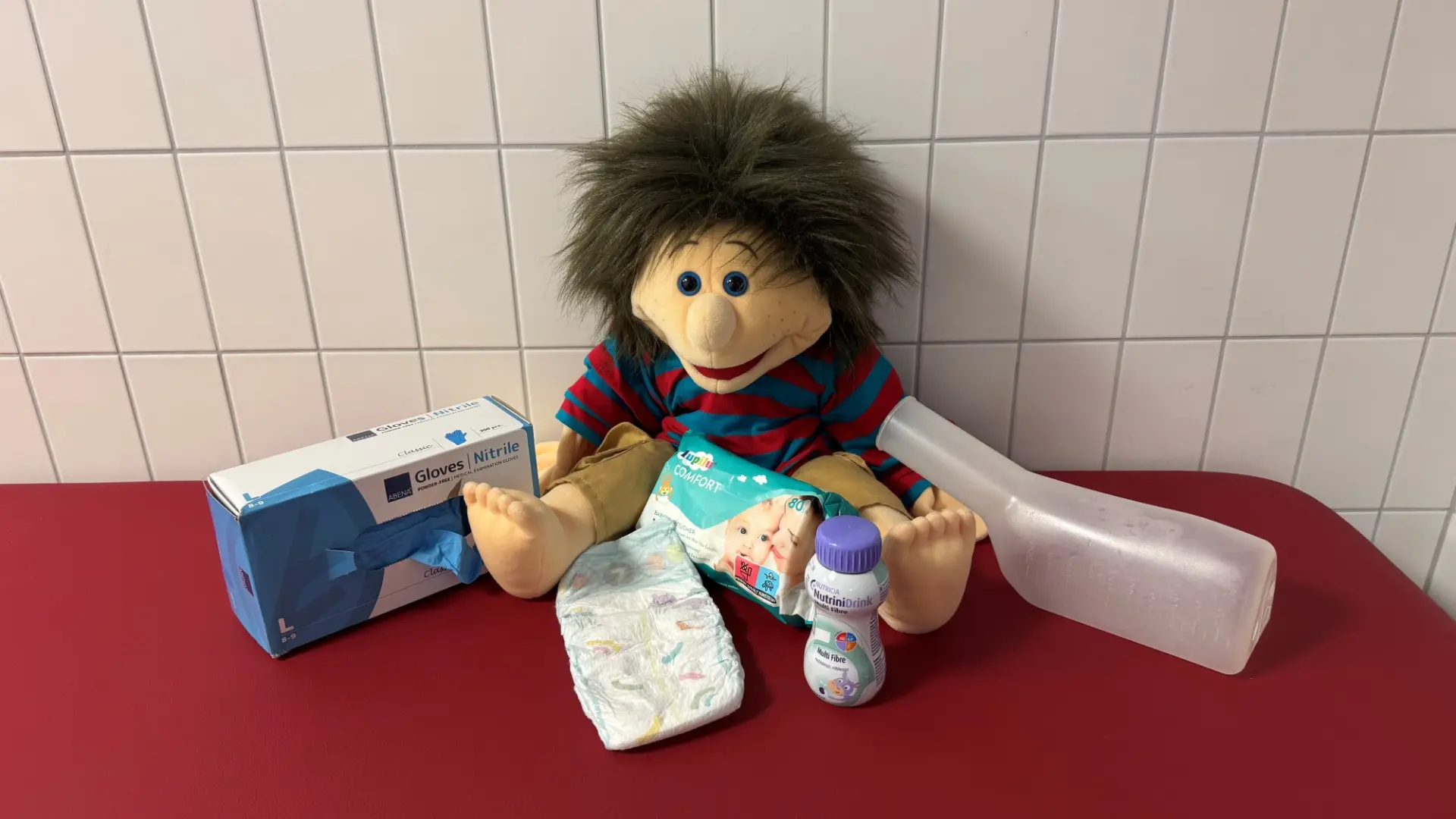 Pflegeutensilien wie Einmalhandschuhe, Feuchttücher, Urinflasche neben einer Handpuppe auf einer roten Liege