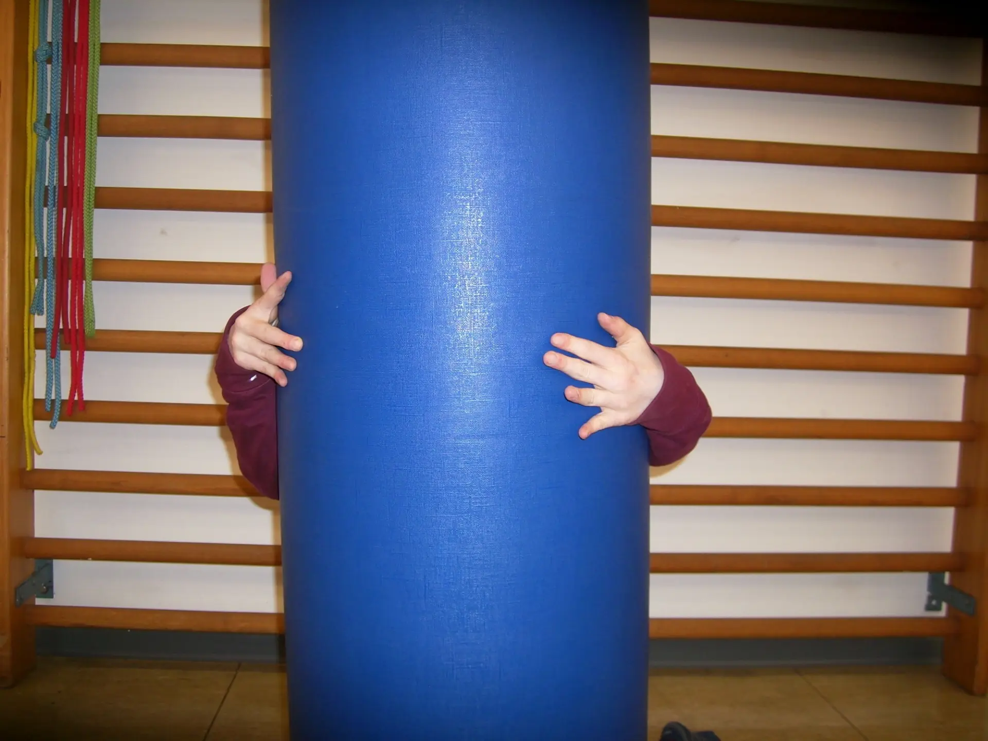 In einer Turnhalle umfasst ein Kind von hinten eine Schaumstoffrolle, die größer ist als es selbst