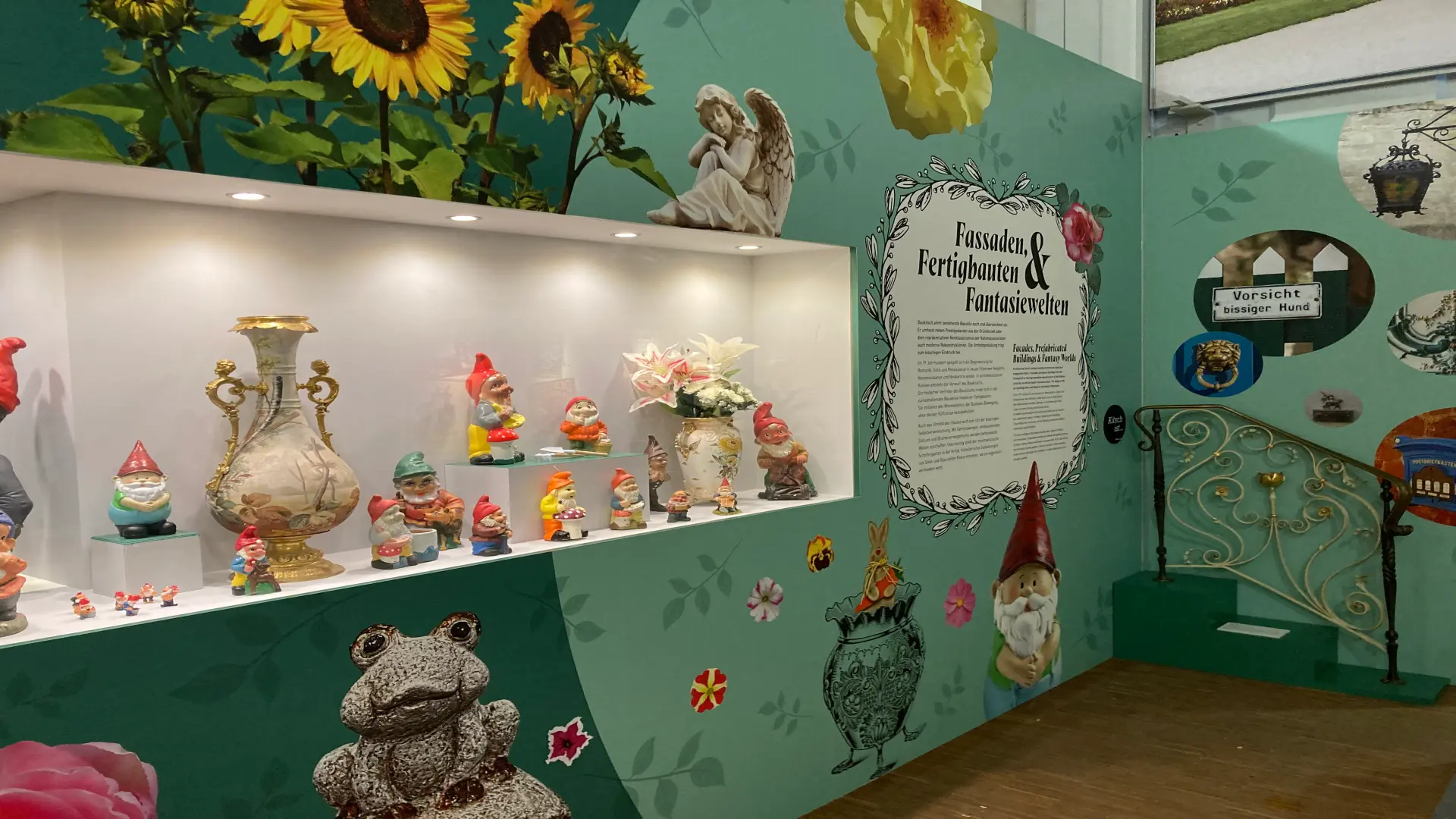Foto: Museumsvitrine mit Gartenzwergen und dekorativen Objekten in einer Ausstellung. Die Wände sind mit Motiven wie Sonnenblumen, Blumen, einem Engel, einem Frosch und weiteren Zierelementen gestaltet.