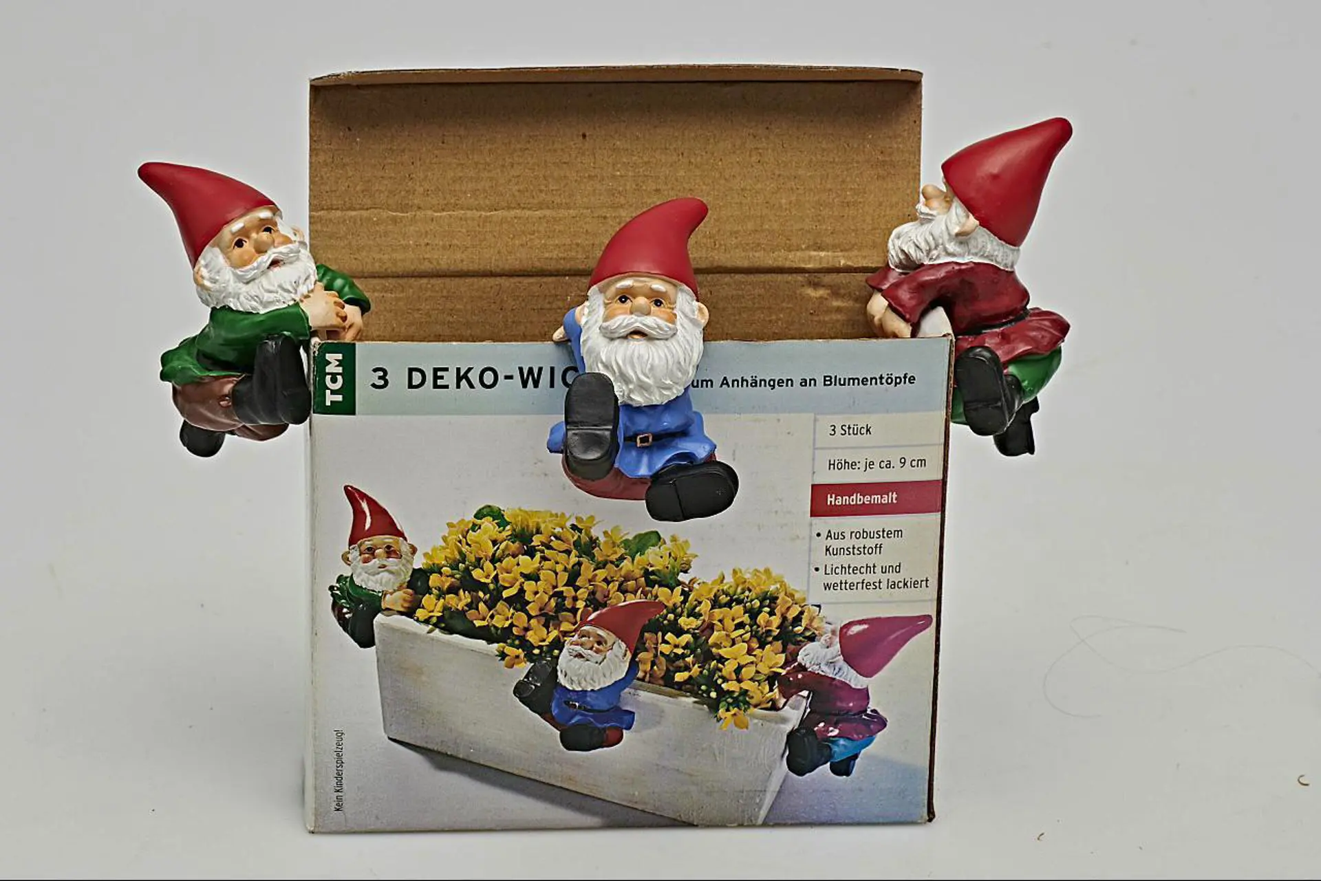 Foto: Drei dekorative Gartenzwerge sitzen auf der Verpackung eines Blumenkastendekor-Sets und präsentieren die beworbenen Figuren.