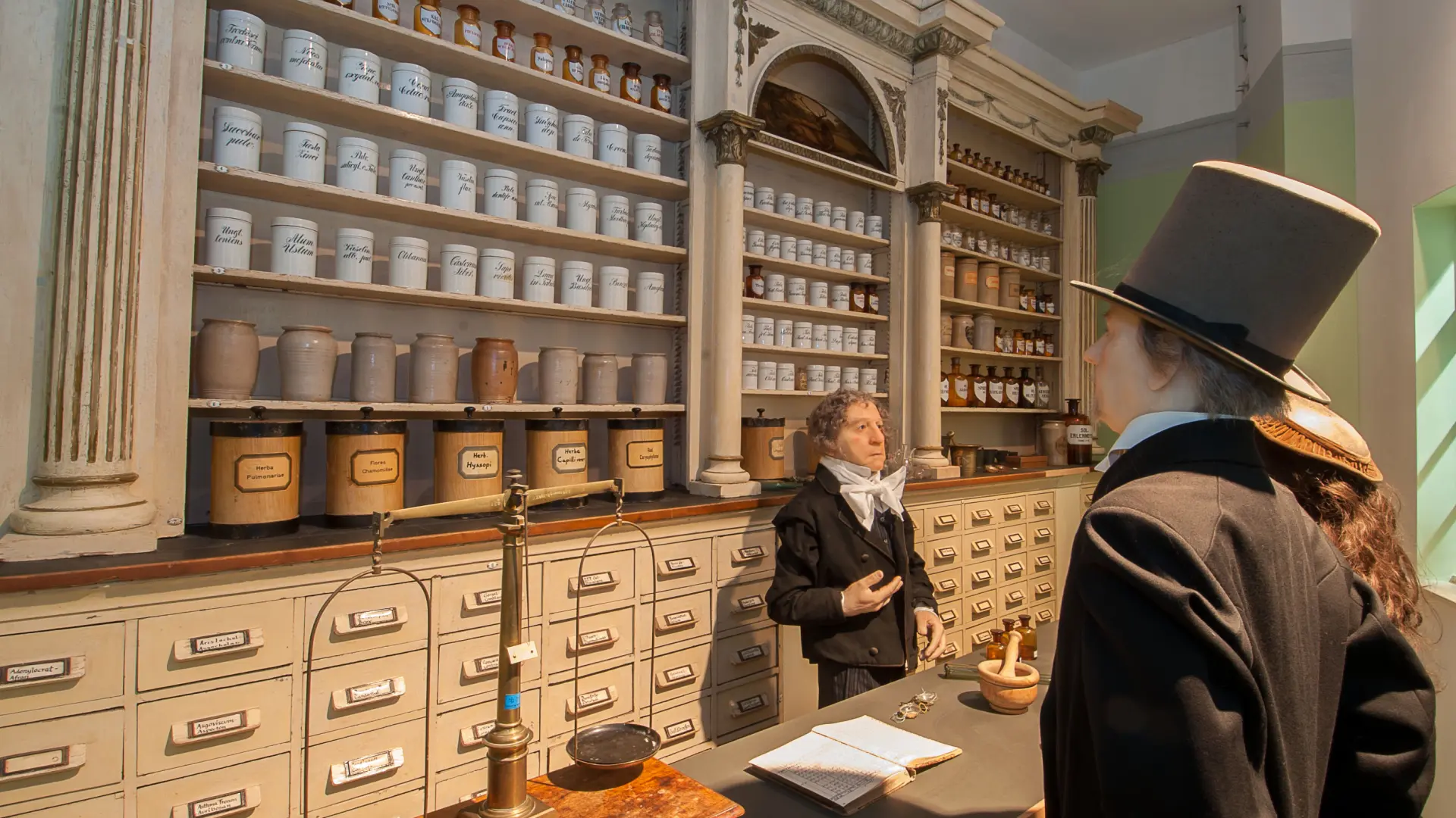 Historische Apothekeneinrichtung mit hohen Regalen voller beschrifteter Arzneigefäße und Schubladen. Zwei Personen in zeitgenössischer Kleidung des 19. Jahrhunderts stehen am Verkaufstresen
