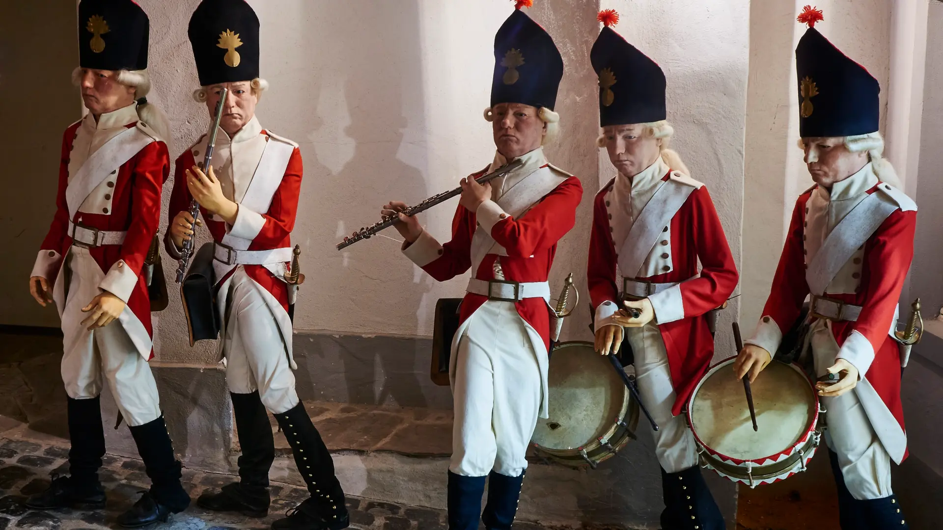 Foto: Szene aus der Ausstellung mit fünf Figuren einer historischen Militärkapelle in roten Uniformen mit Trommeln und Flöten.