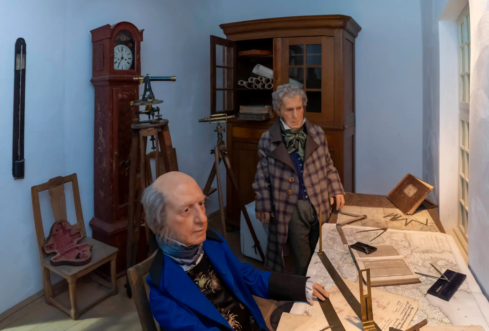 Foto: Szene aus der Ausstellung mit einem Büro mit Vermessungsinstrumenten, Karten und zwei Figuren in zeitgenössischer Kleidung.