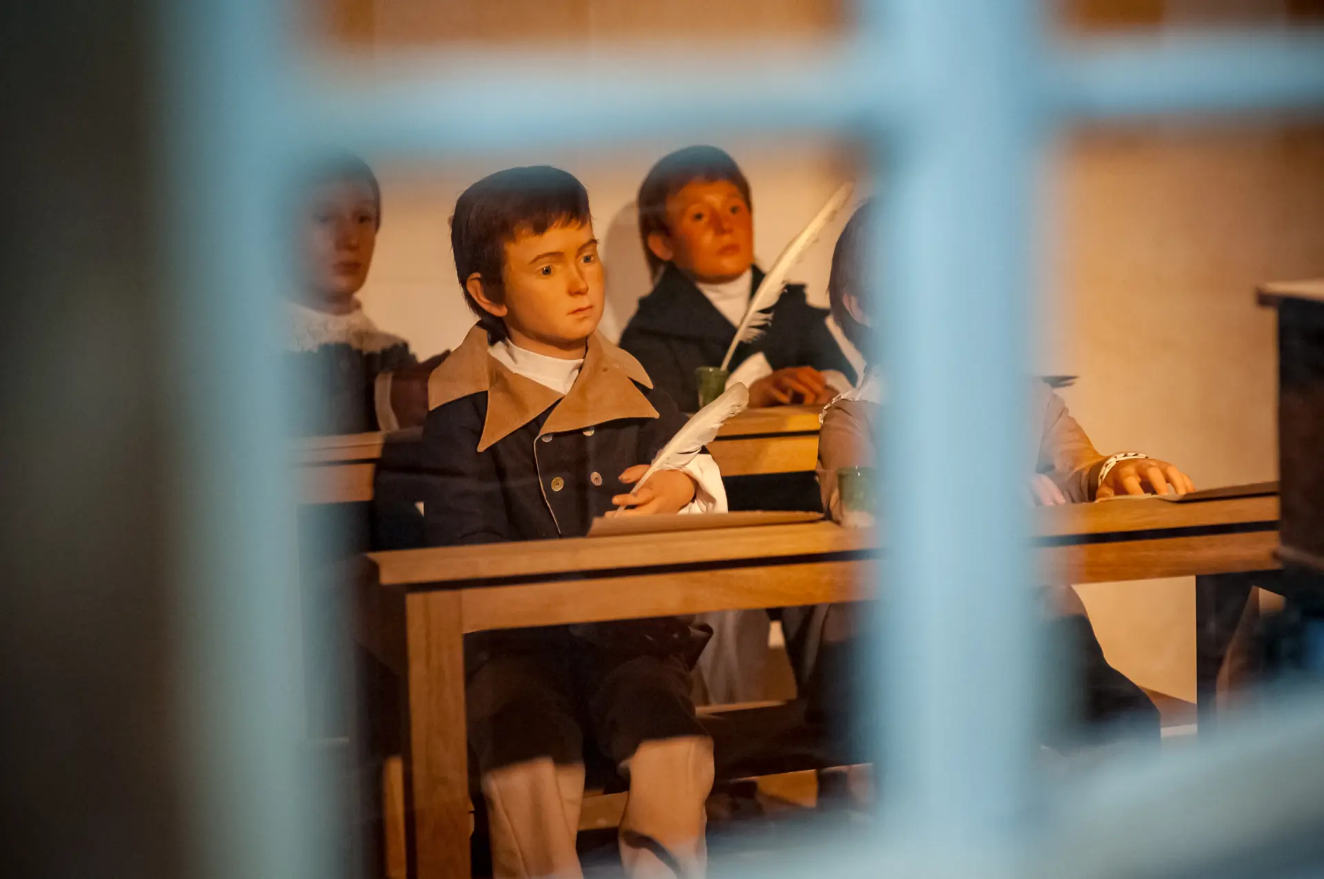 Foto:Szene aus der Ausstellung mit Klassenzimmer mit sitzenden Schulkindern in zeitgenössischer Kleidung.