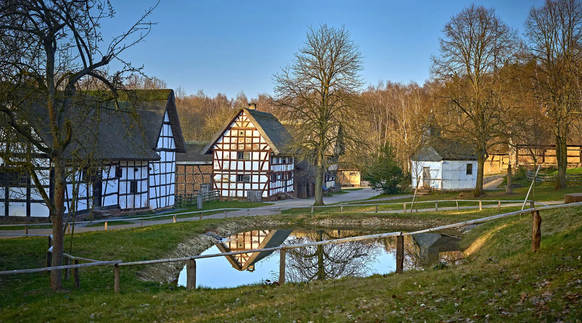 Das Bild zeigt einen Teich hinter einem simplen Zaun. Im Hintergrund sind zwei Fachwerkhäuser und eine weiße Fachwerkkapelle zu sehen.