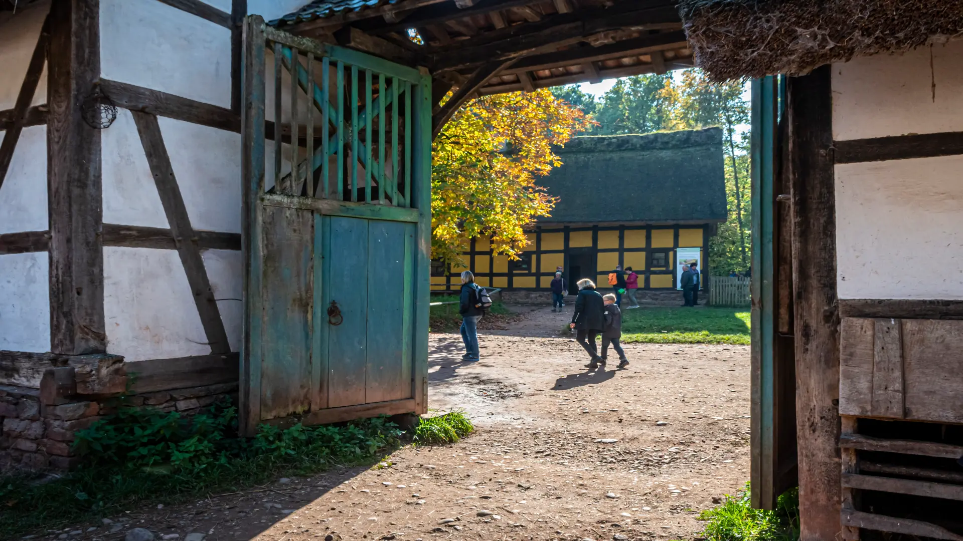 historischer Innenhof mit mehreren Fachwerkgebäuden zu sehen. Im Vordergrund steht ein großes, geöffnetes Holztor mit grün gestrichenen Teilen. Links und rechts begrenzen Fachwerkwände den Durchgang. Im Hintergrund befindet sich ein weiteres Fachwerkhaus mit gelben Gefachen und einem dunklen Reetdach.