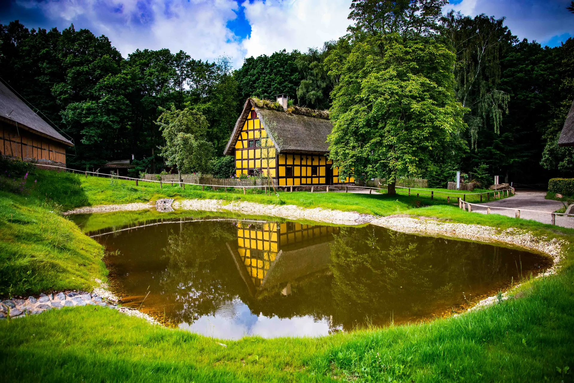 Das Foto zeigt ein gelbes Fachwerkhaus hinter einem großen Teich im Vordergrund. Neben dem Fachwerkhaus steht ein Baum.
