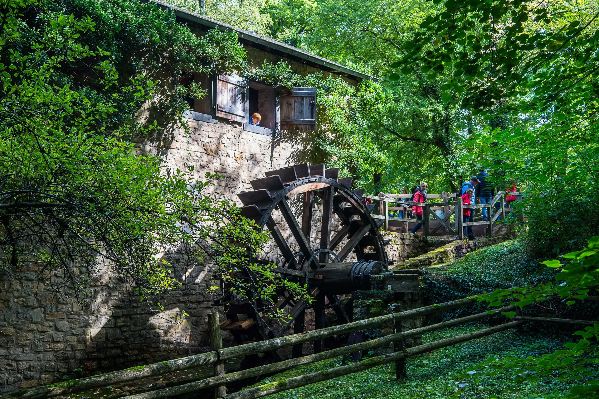 Wassermühle im Wald. Die Mühle ist ein Steingebäude mit einem Fenster im Obergeschoss, aus dem ein Junge heraus schaut. Darunter ist ein großes Wasserrad. Weiter oben am Hang laufen Menschen hinter einem Zaun.