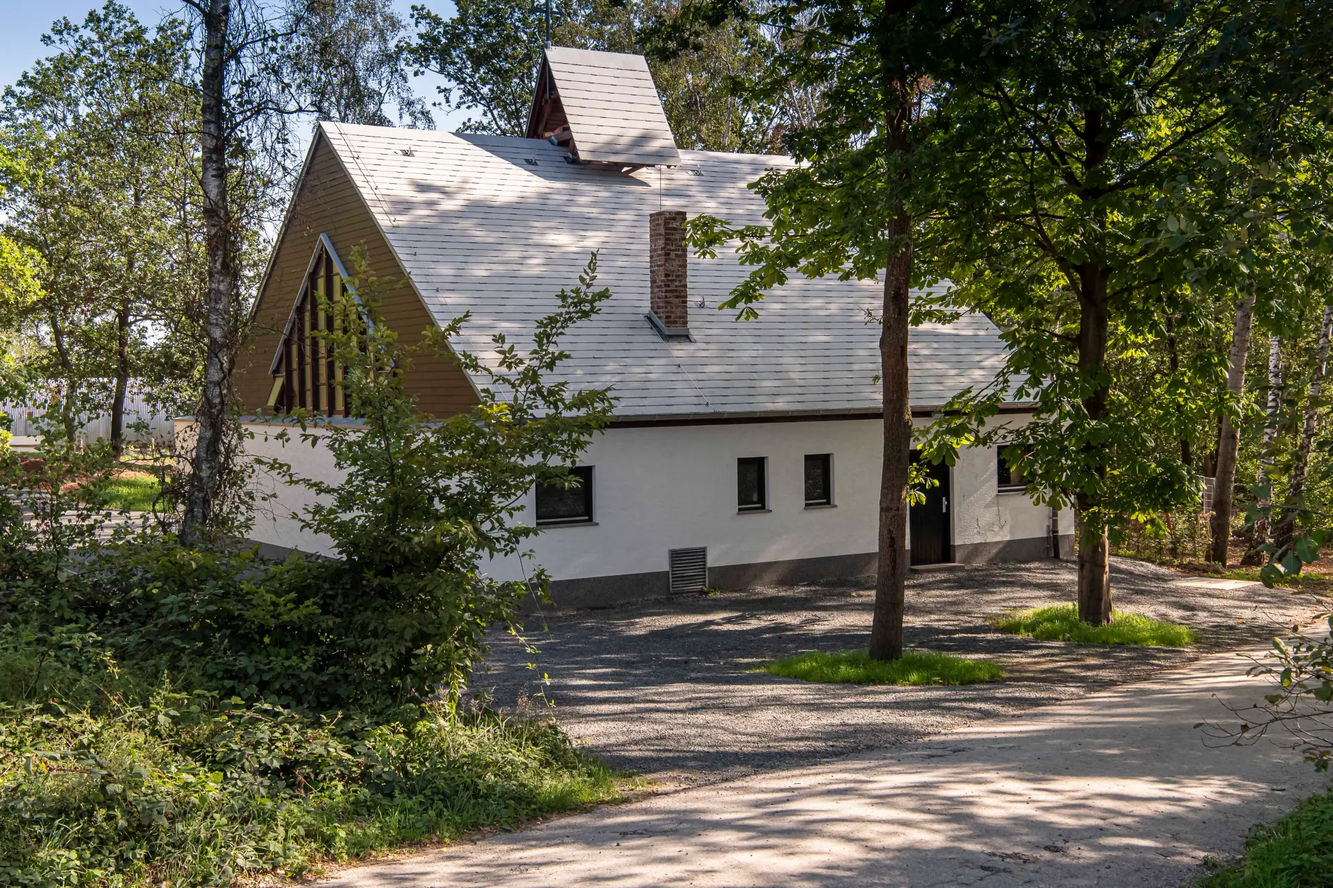 Das Bild zeigt eine weiße Kapelle aus der Nachkriegszeit, umgeben von Bäumen.
