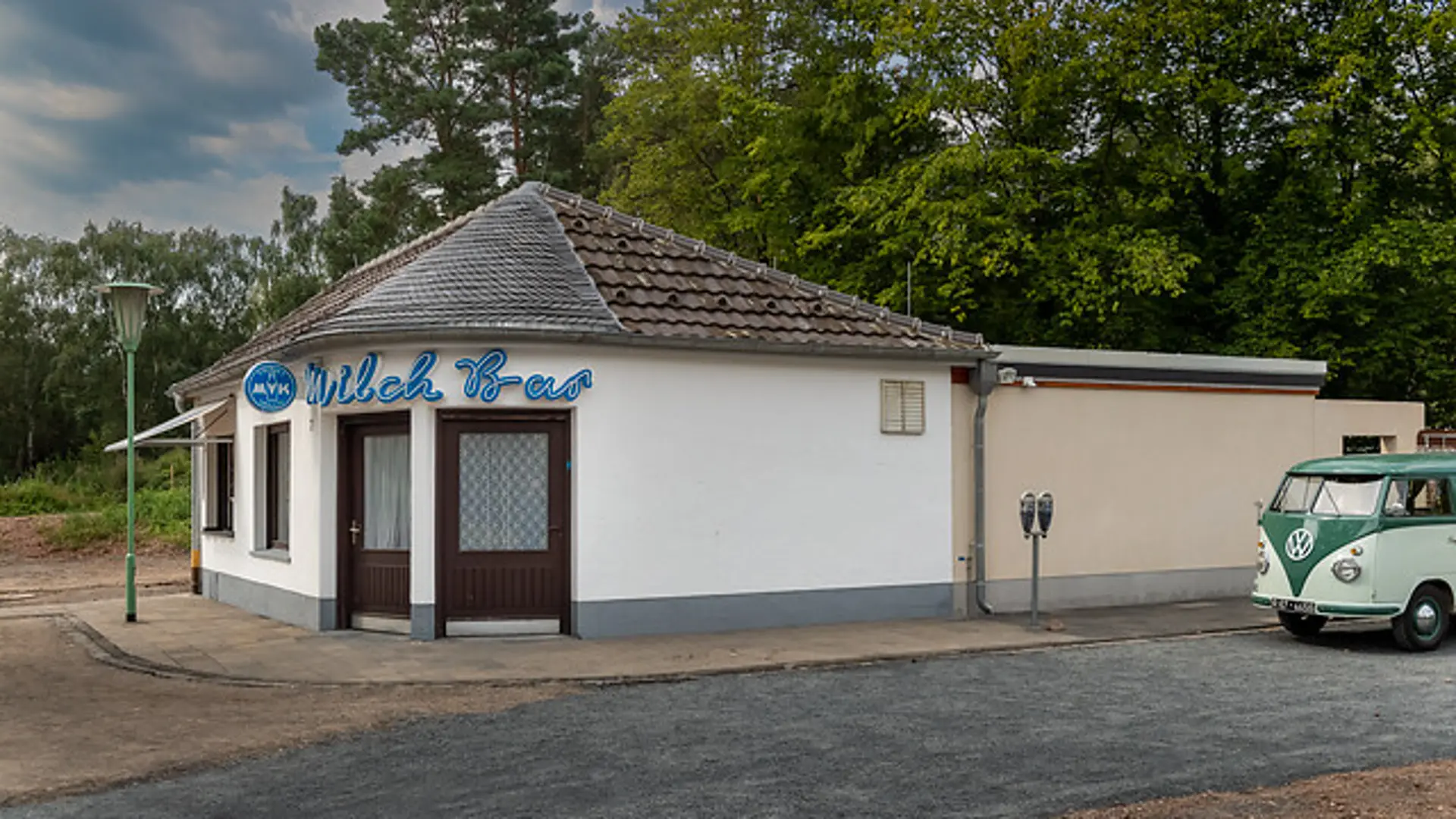 einstöckiges weißes Gebäude mit grauem Dach. Das Gebäude geht über Eck und hat zwei Eingangstüren in beide Richtungen an der Ecke. Über dem Eingang steht Milch-Bar in Leuchtschrift.