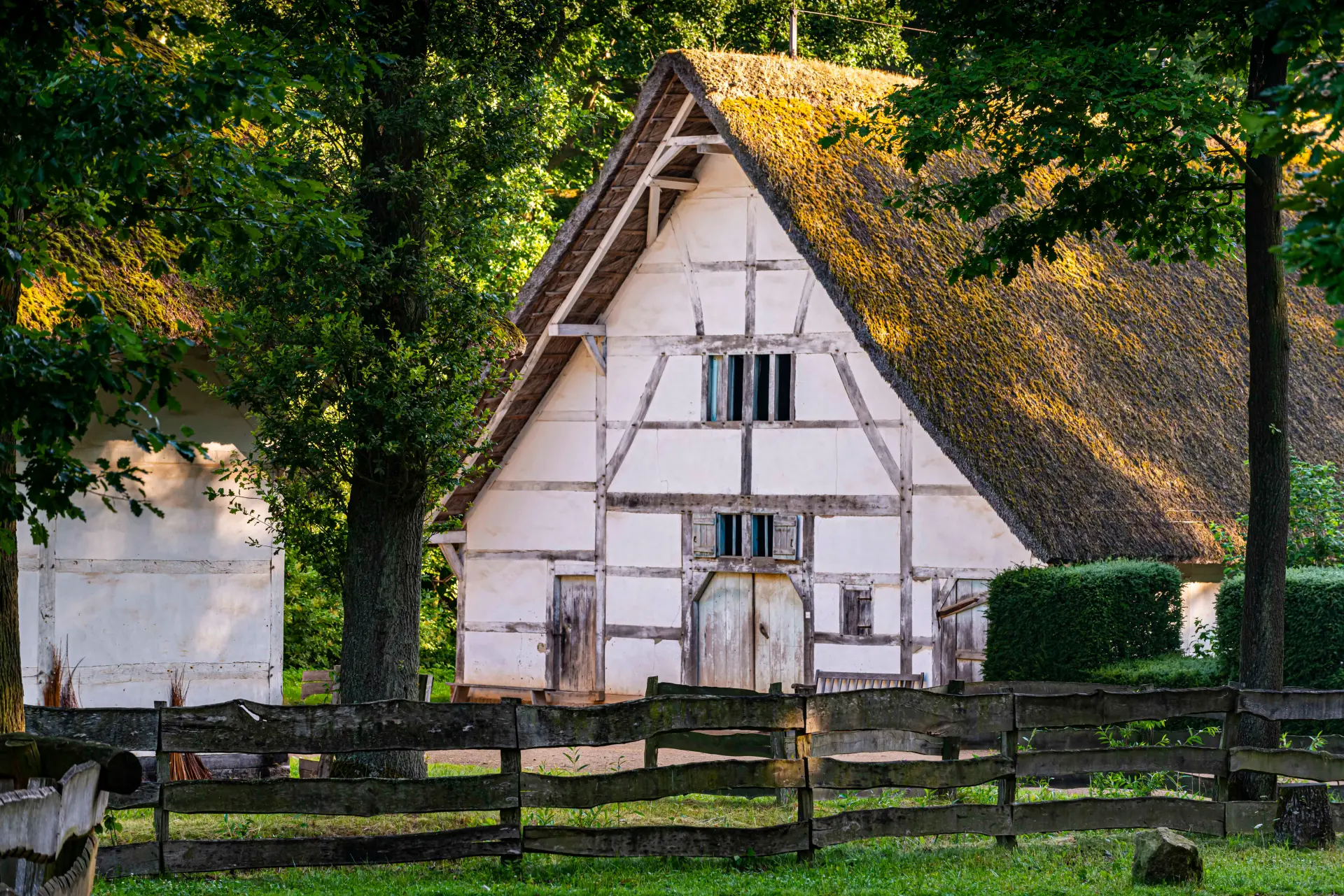 Fachwerkhaus mit Strohdach hinter Holzzaun und Bäumen.
