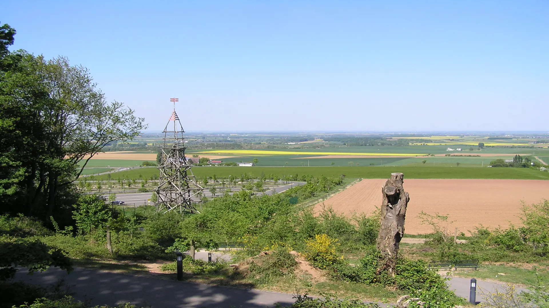 Weite Landschaft mit Feldern und Wiesen im Hintergrund, davor ein Parkplatzbereich mit Wegen und Zäunen. Rechts steht eine große hölzerne Turm- oder Gerüstkonstruktion, während der Blick über die offene ländliche Umgebung bis zum Horizont reicht.