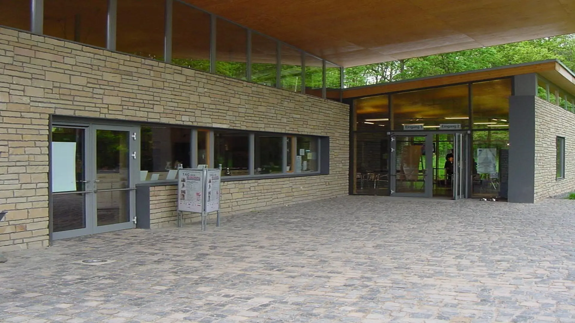 Moderner Eingangsbereich des Freilichtmuseums mit heller Natursteinfassade, großen Glasflächen und überdachtem Zugang. Vor dem Gebäude führt ein gepflasterter Vorplatz zu den Eingängen,