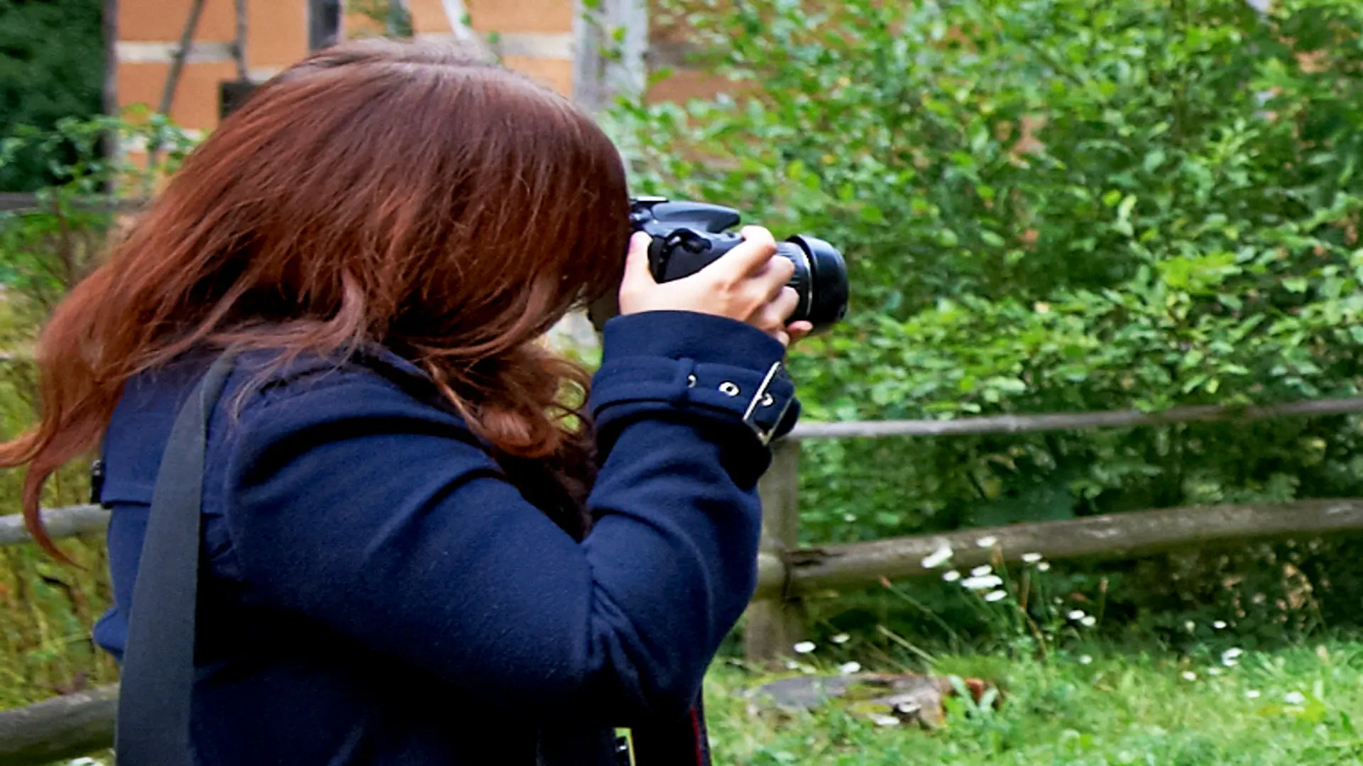 Eine Person mit langen roten Haaren fotografiert mit einer DSLR-Kamera in einem grünen Garten mit Büschen und einem Holzzaun im Hintergrund.