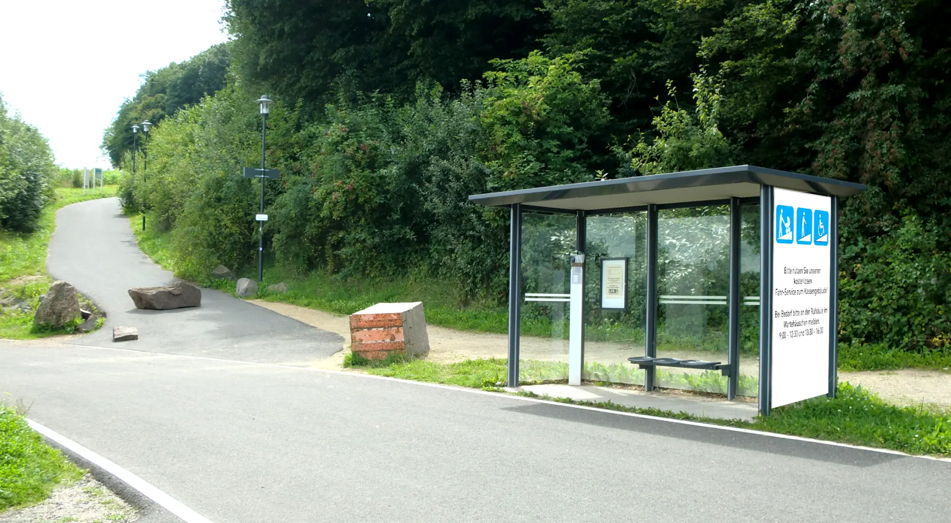 Bushaltestelle bzw. Fahrservice-Haltestelle am Rand eines Waldgebiets mit modernem Wartehäuschen aus Glas und Sitzbank. Ein asphaltierter Weg führt durch die grüne Umgebung, gesäumt von Bäumen und Sträuchern.
