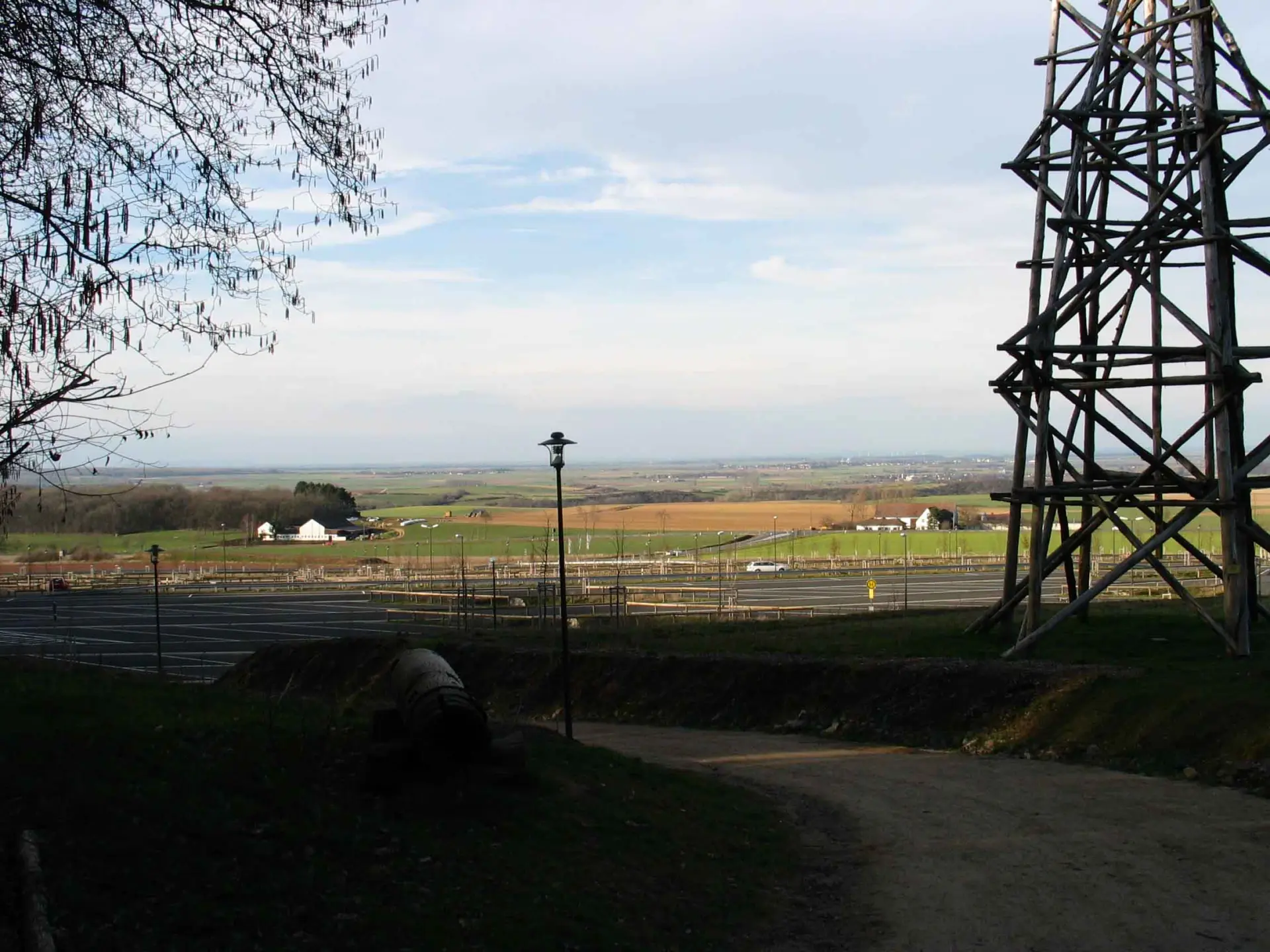 Weite Landschaft mit Feldern und Wiesen im Hintergrund, davor ein Parkplatzbereich mit Wegen und Zäunen. Rechts steht eine große hölzerne Turm- oder Gerüstkonstruktion, während der Blick über die offene ländliche Umgebung bis zum Horizont reicht.