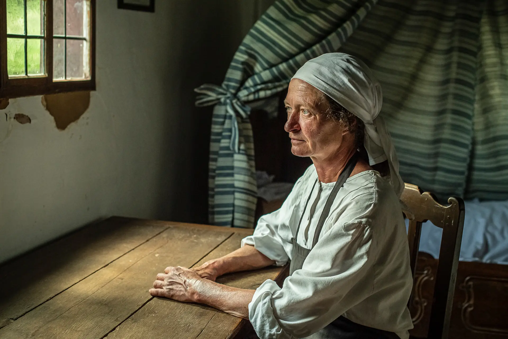 Eine Frau mit weißem Kopftuch in weißer Bluse sitzt an einem Holztisch.