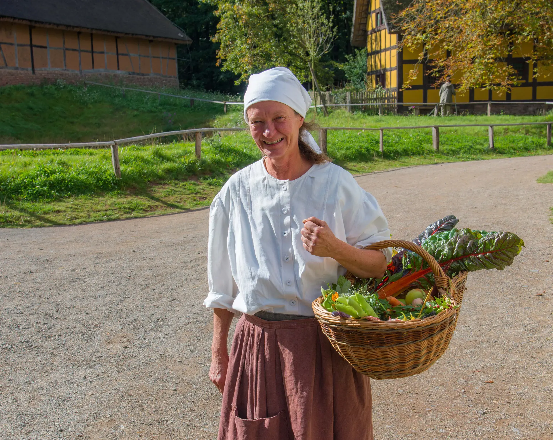 Eine Frau mit weißer Bluse, Kopftuch und Schürze mit einem Korb voller Gemüse am Arm.