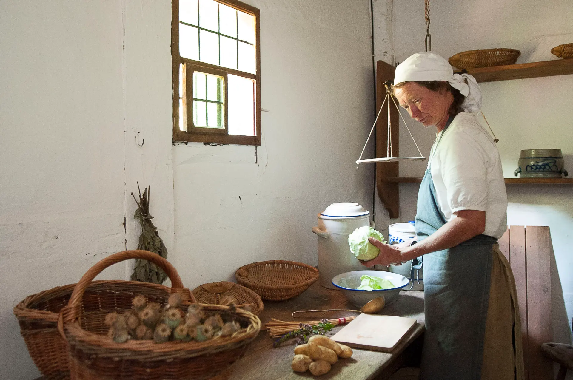 Eine Frau mit Schürze und weißem Kopftuch steht in einer Küche mit Holztisch.