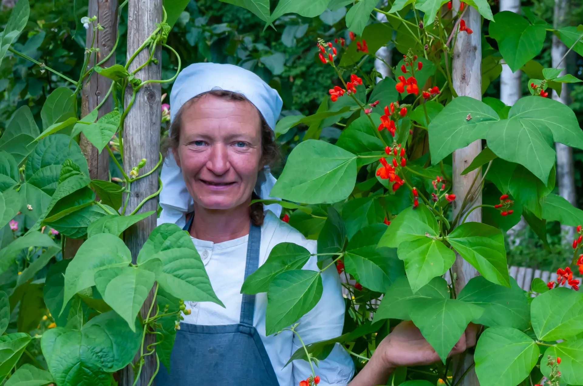 Eine Frau mit weißem Kopftuch schaut zwischen rankenden Blumen hervor.