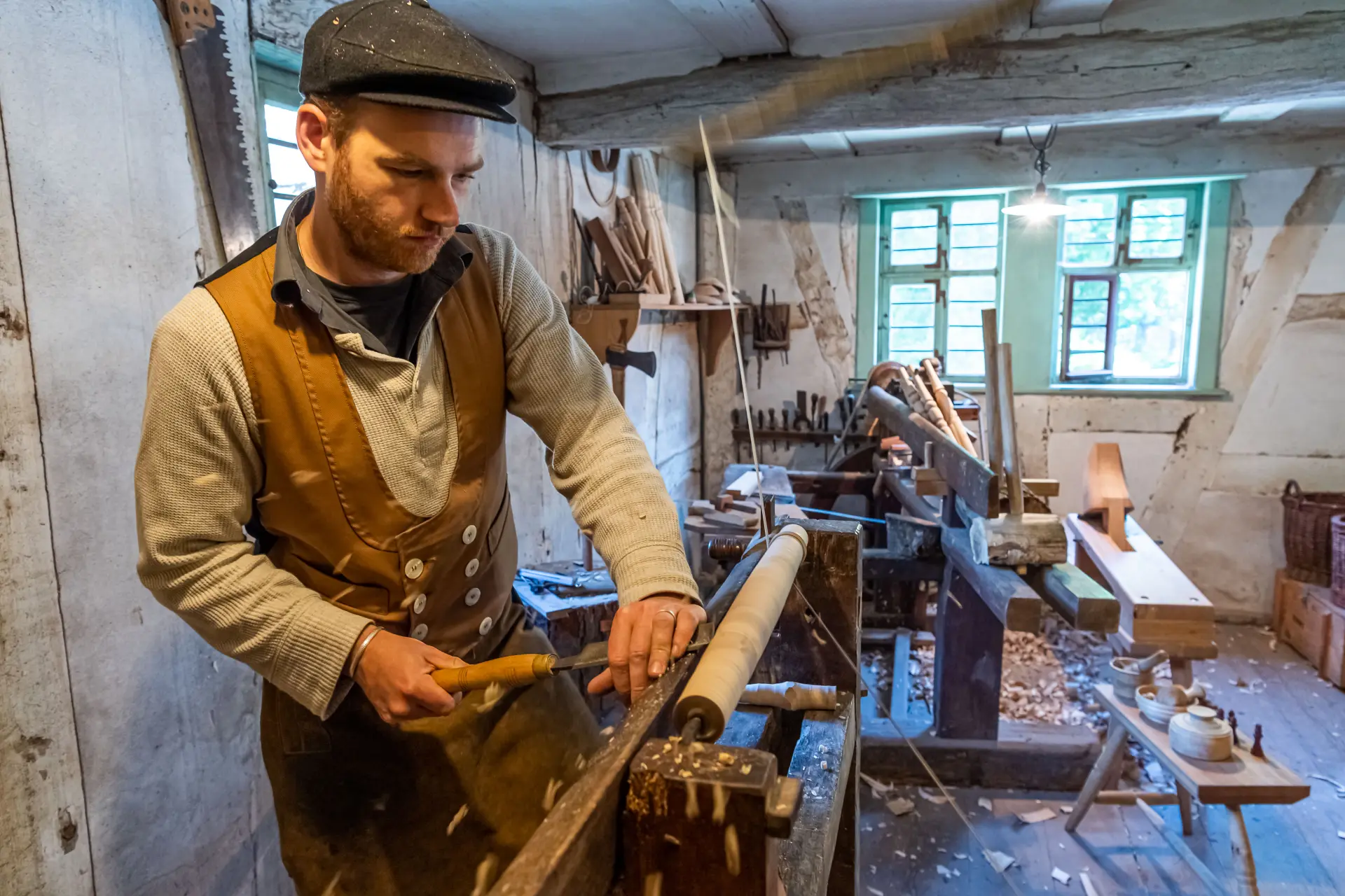 Eine ältere Frau sitzt an einem rustikalen Holztisch in einem historischen, steinernen Raum mit warmem Licht. Sie trägt runde Brille, ein graues Kopftuch, einen dunklen Schal und schlichte, traditionelle Kleidung. In ihren Händen hält sie einen drahtförmigen Gegenstand, der wie eine handgefertigte Mausefalle aussieht. Auf dem Tisch liegen weitere Drahtkonstruktionen sowie Werkzeuge wie Zangen.