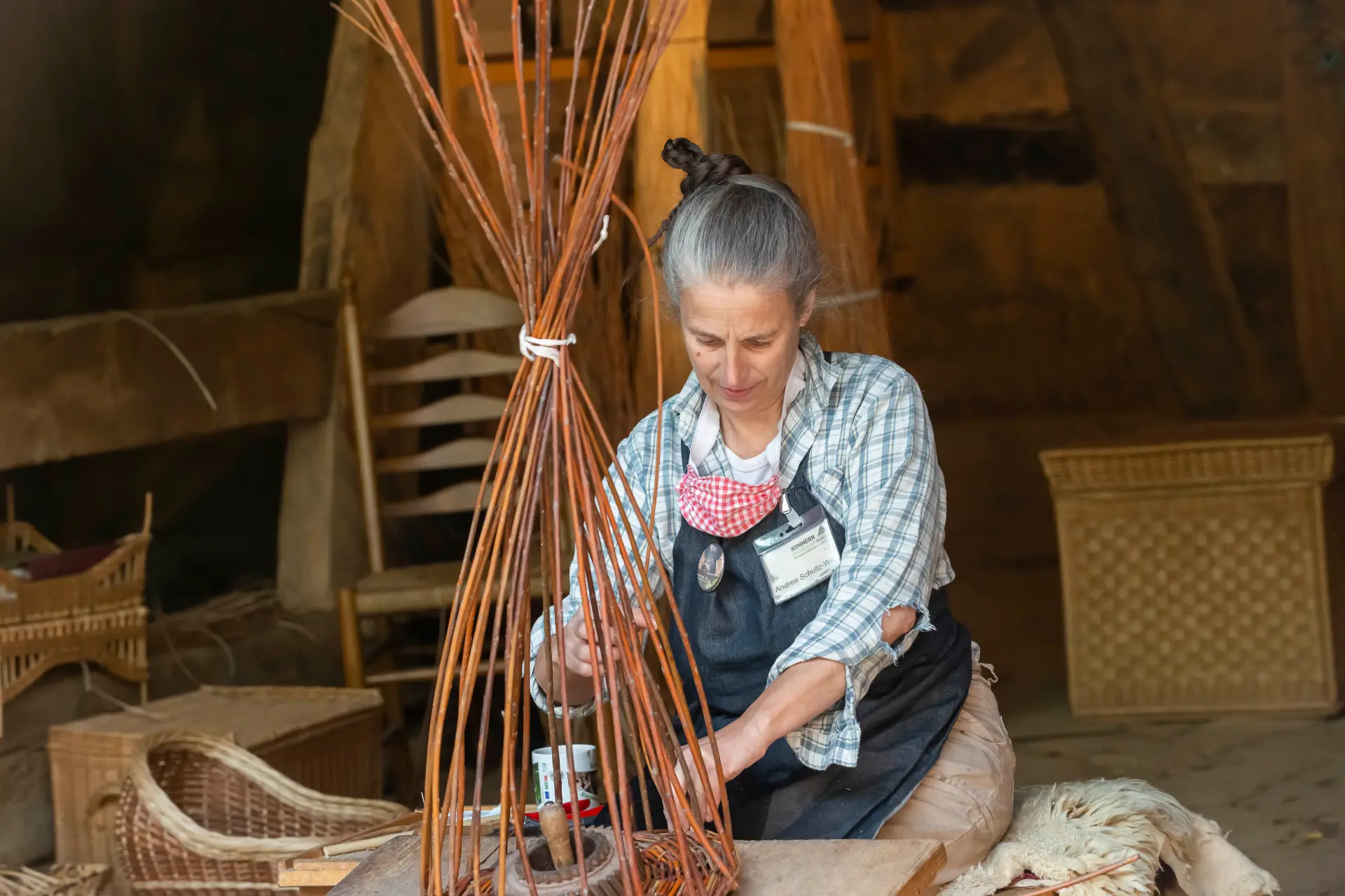 Eine ältere Frau sitzt an einem rustikalen Holztisch in einem historischen, steinernen Raum mit warmem Licht. Sie trägt runde Brille, ein graues Kopftuch, einen dunklen Schal und schlichte, traditionelle Kleidung. In ihren Händen hält sie einen drahtförmigen Gegenstand, der wie eine handgefertigte Mausefalle aussieht. Auf dem Tisch liegen weitere Drahtkonstruktionen sowie Werkzeuge wie Zangen.