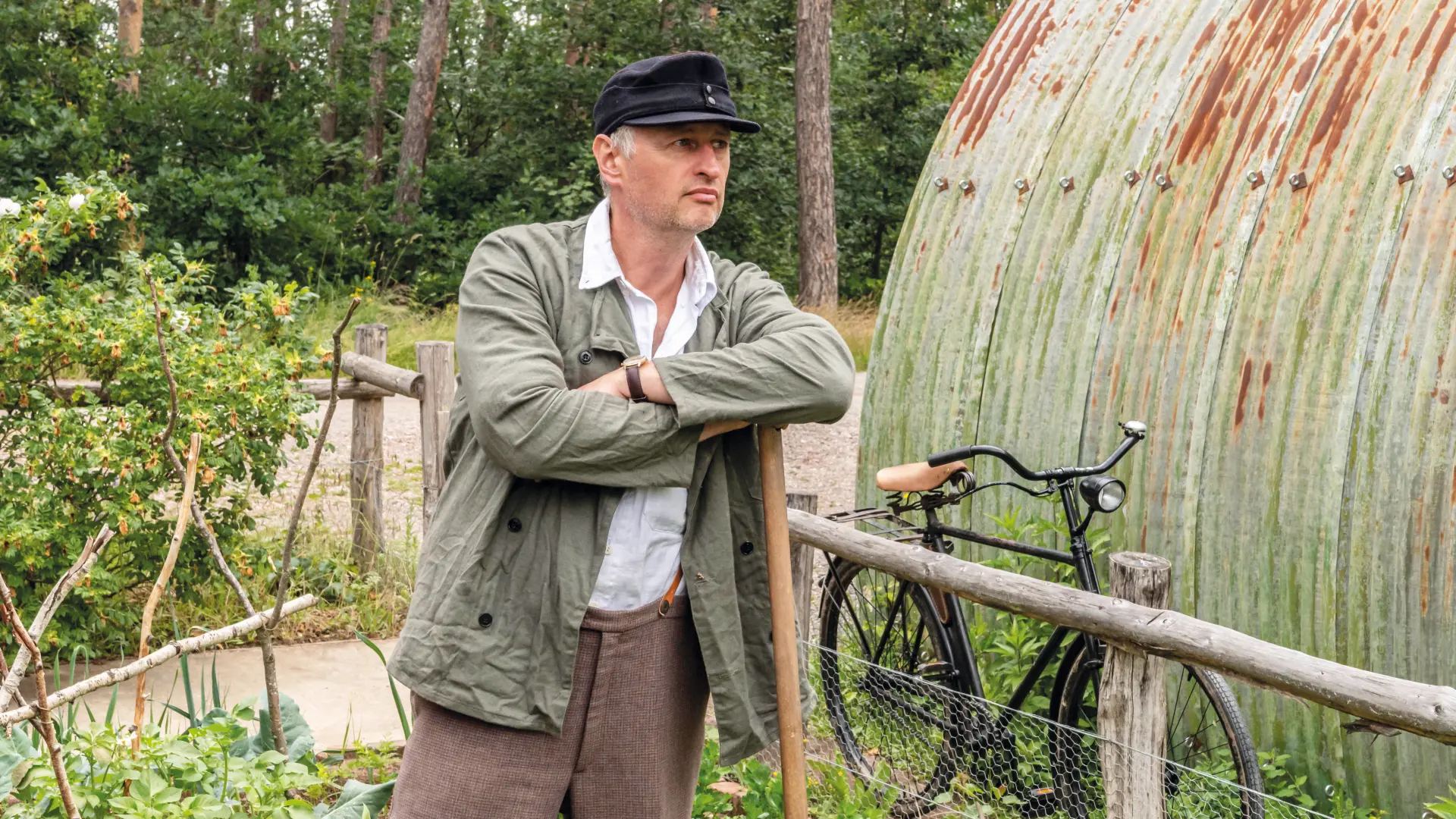 Mann in brauner Hose und grüner Jacke mit Hut stützt sich auf eine Schaufel in einem Garten.