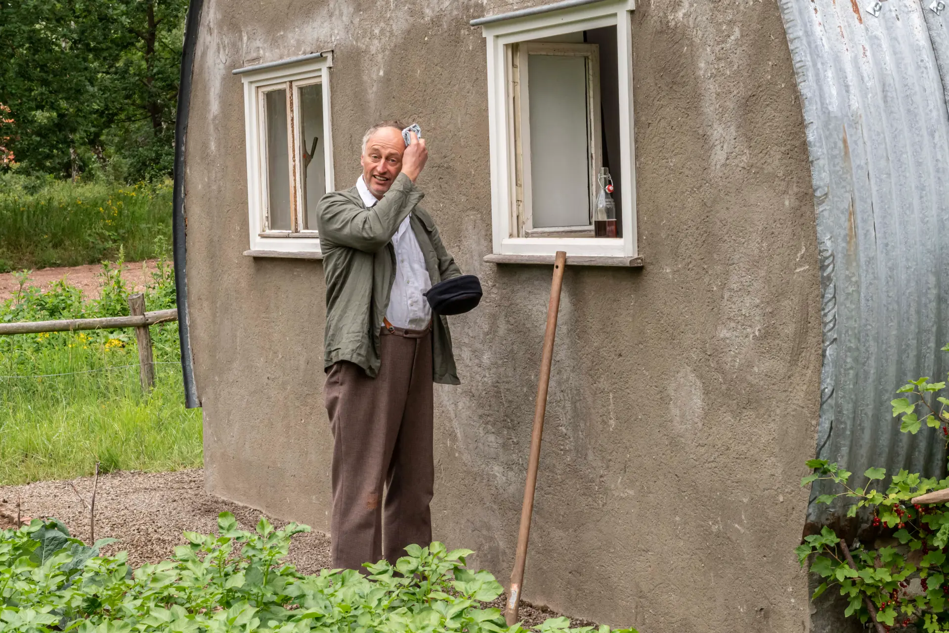 Kriegsheimkehrer steht vor der Nissenhütte, eine braune Fassade und weiße Fenster. Er wischt sich den Schweiß mit einem Taschentuch von der Stirn.