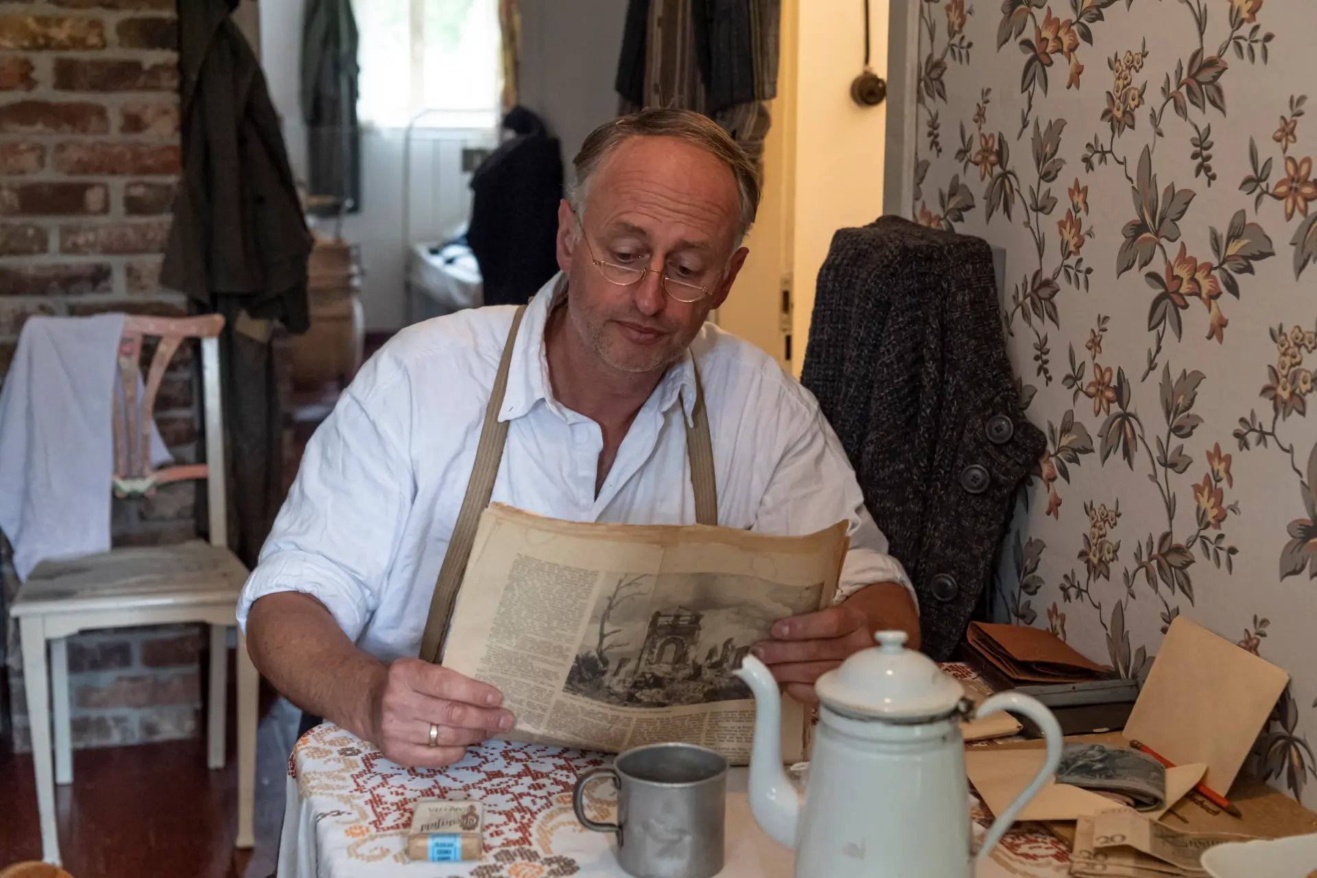 Kriegsheimkehrer sitzt am Küchentisch vor einer Blümchentapete und liest eine Zeitung.