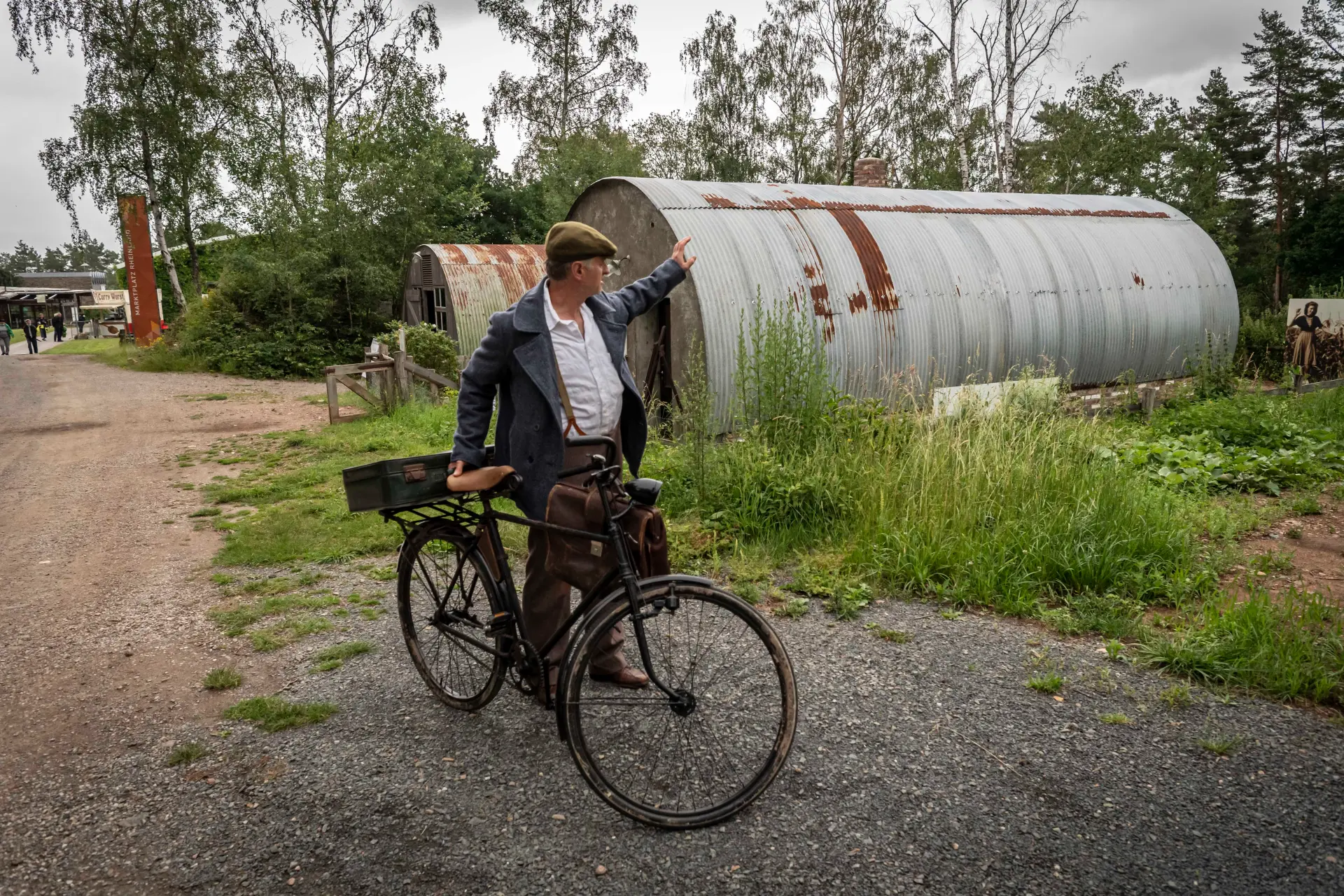 Kriegsheimkehrer steht mit seinem Fahrrad auf einem Schotterweg. Im Hintergrund ist das Blech der Nissenhütten zu sehen.