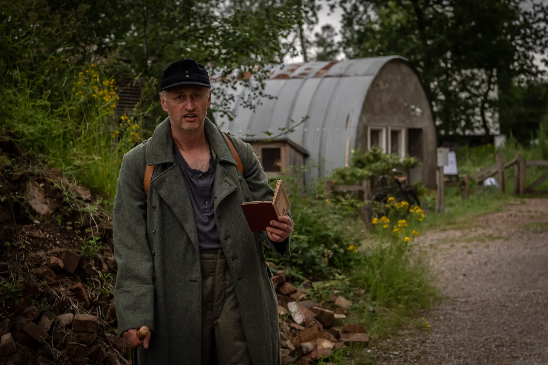 Der Kriegsheimkehrer steht vor Trümmerschutt und der Nissenhütte und liest aus einem kleinen Büchlein vor, das er in der Hand hält.