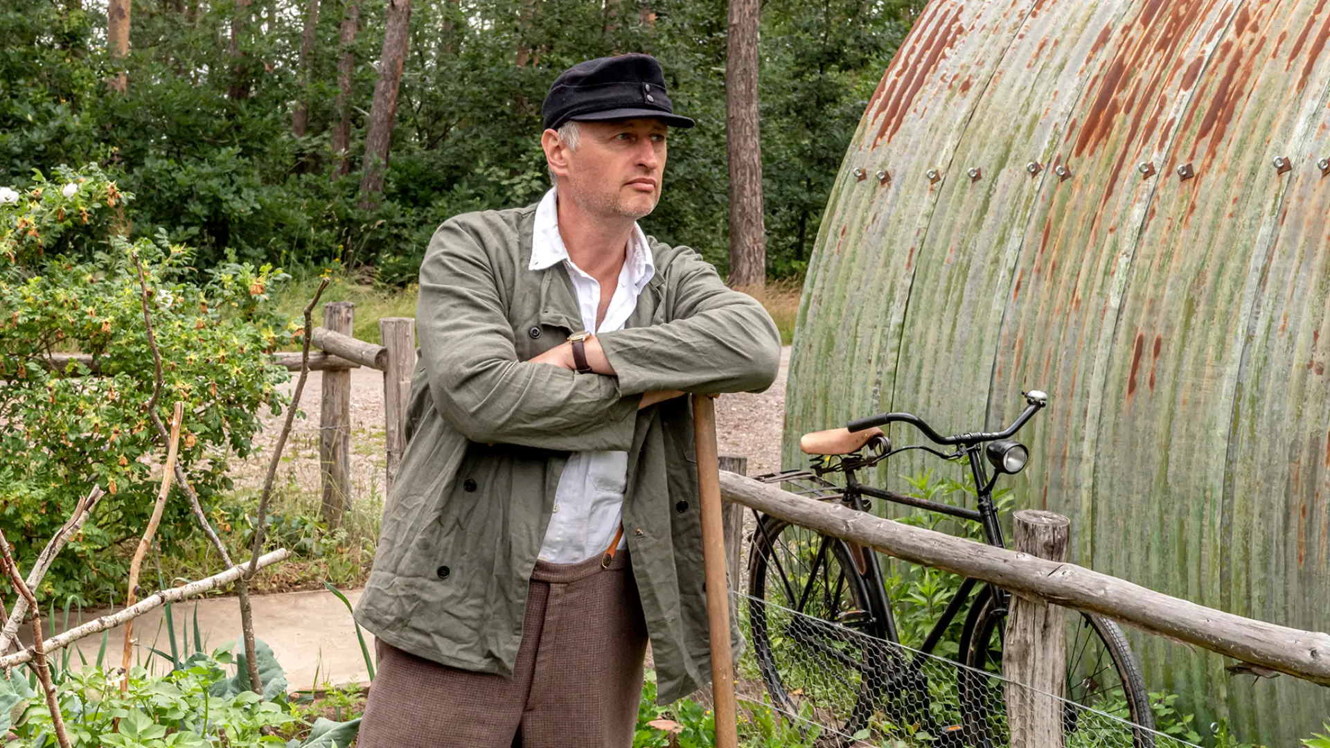 Mann in brauner Hose und grüner Jacke mit Hut stützt sich auf eine Schaufel in einem Garten.