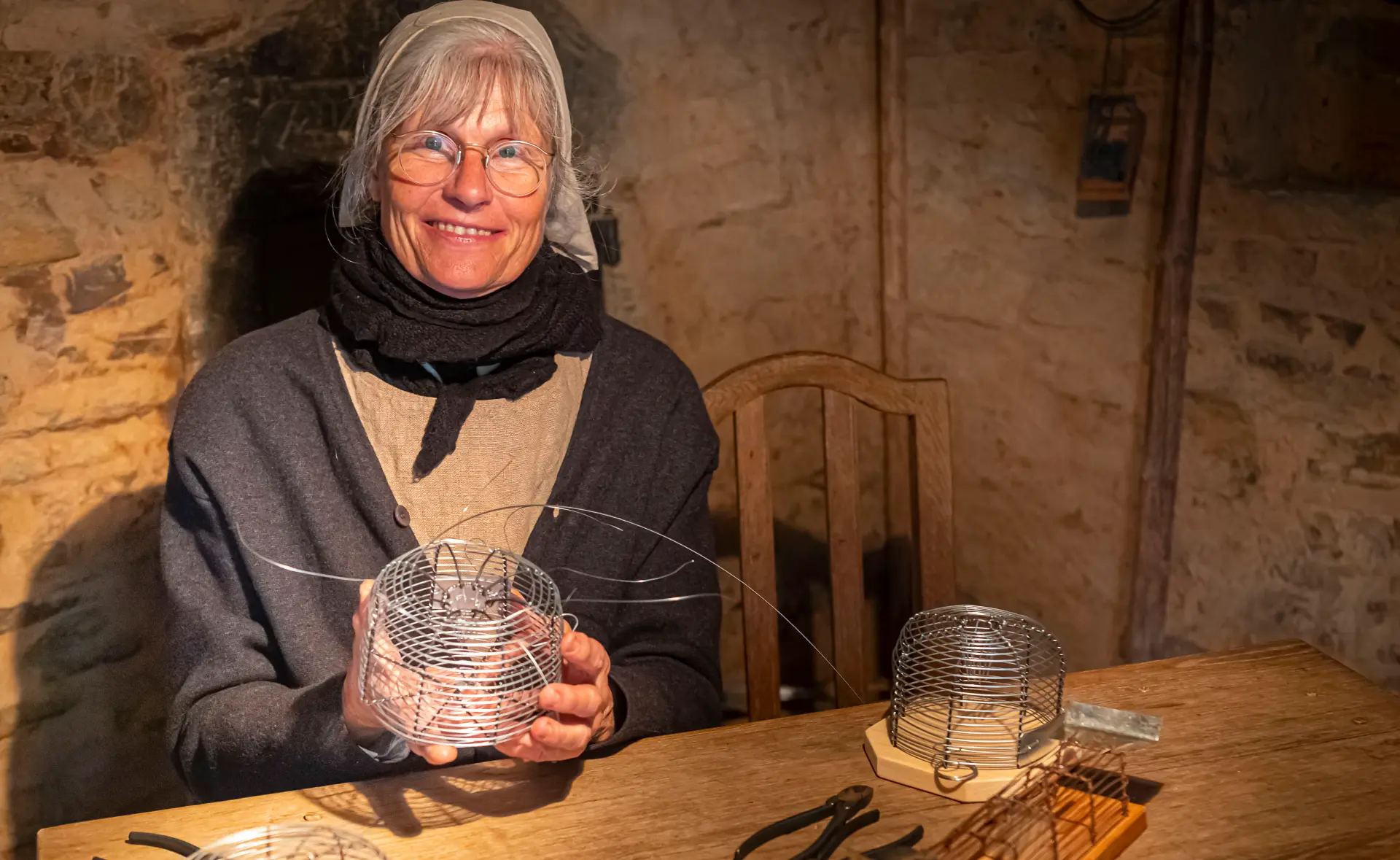 Eine ältere Frau sitzt an einem rustikalen Holztisch in einem historischen, steinernen Raum mit warmem Licht. Sie trägt runde Brille, ein graues Kopftuch, einen dunklen Schal und schlichte, traditionelle Kleidung. In ihren Händen hält sie einen drahtförmigen Gegenstand, der wie eine handgefertigte Mausefalle aussieht. Auf dem Tisch liegen weitere Drahtkonstruktionen sowie Werkzeuge wie Zangen.