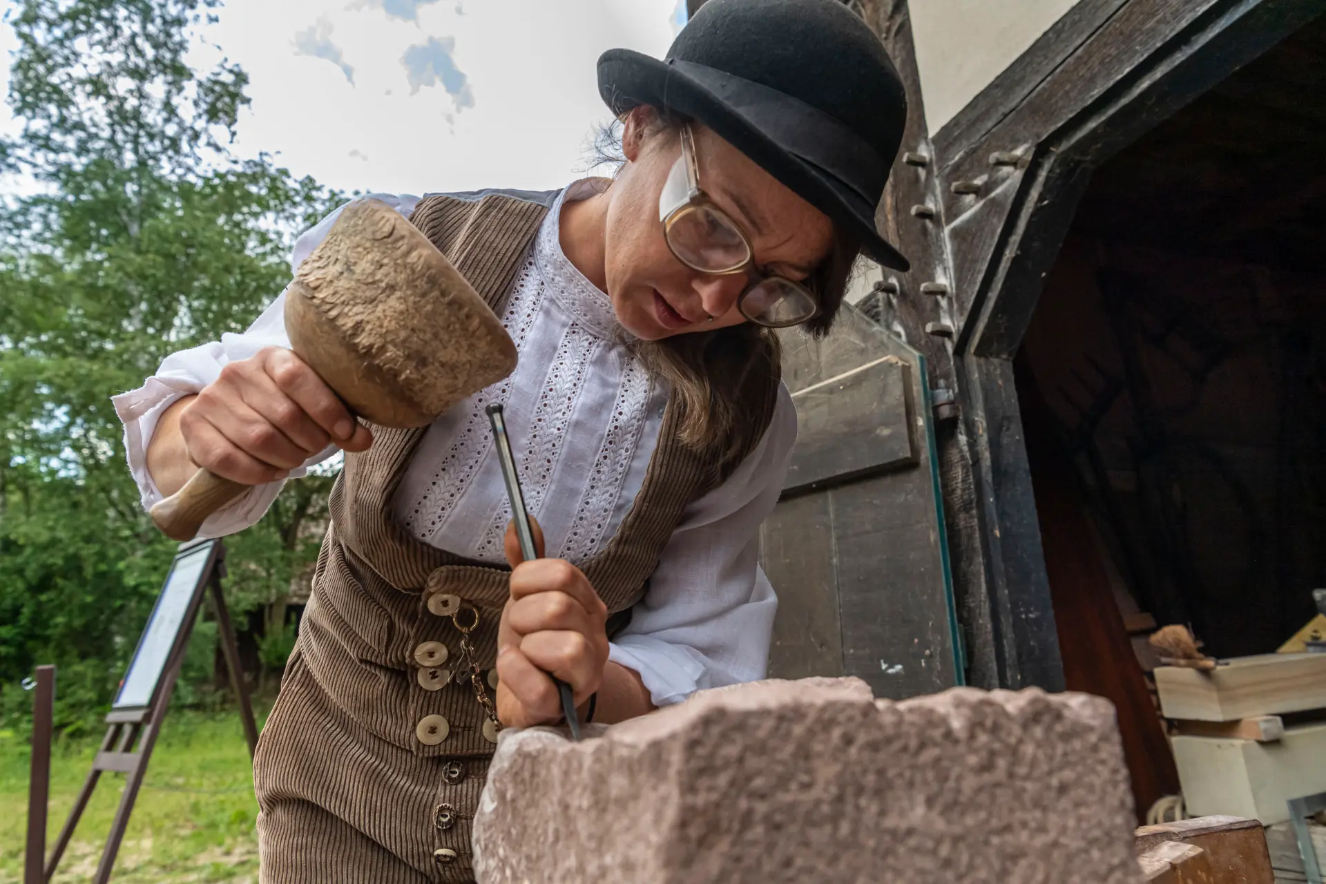 Eine ältere Frau sitzt an einem rustikalen Holztisch in einem historischen, steinernen Raum mit warmem Licht. Sie trägt runde Brille, ein graues Kopftuch, einen dunklen Schal und schlichte, traditionelle Kleidung. In ihren Händen hält sie einen drahtförmigen Gegenstand, der wie eine handgefertigte Mausefalle aussieht. Auf dem Tisch liegen weitere Drahtkonstruktionen sowie Werkzeuge wie Zangen.
