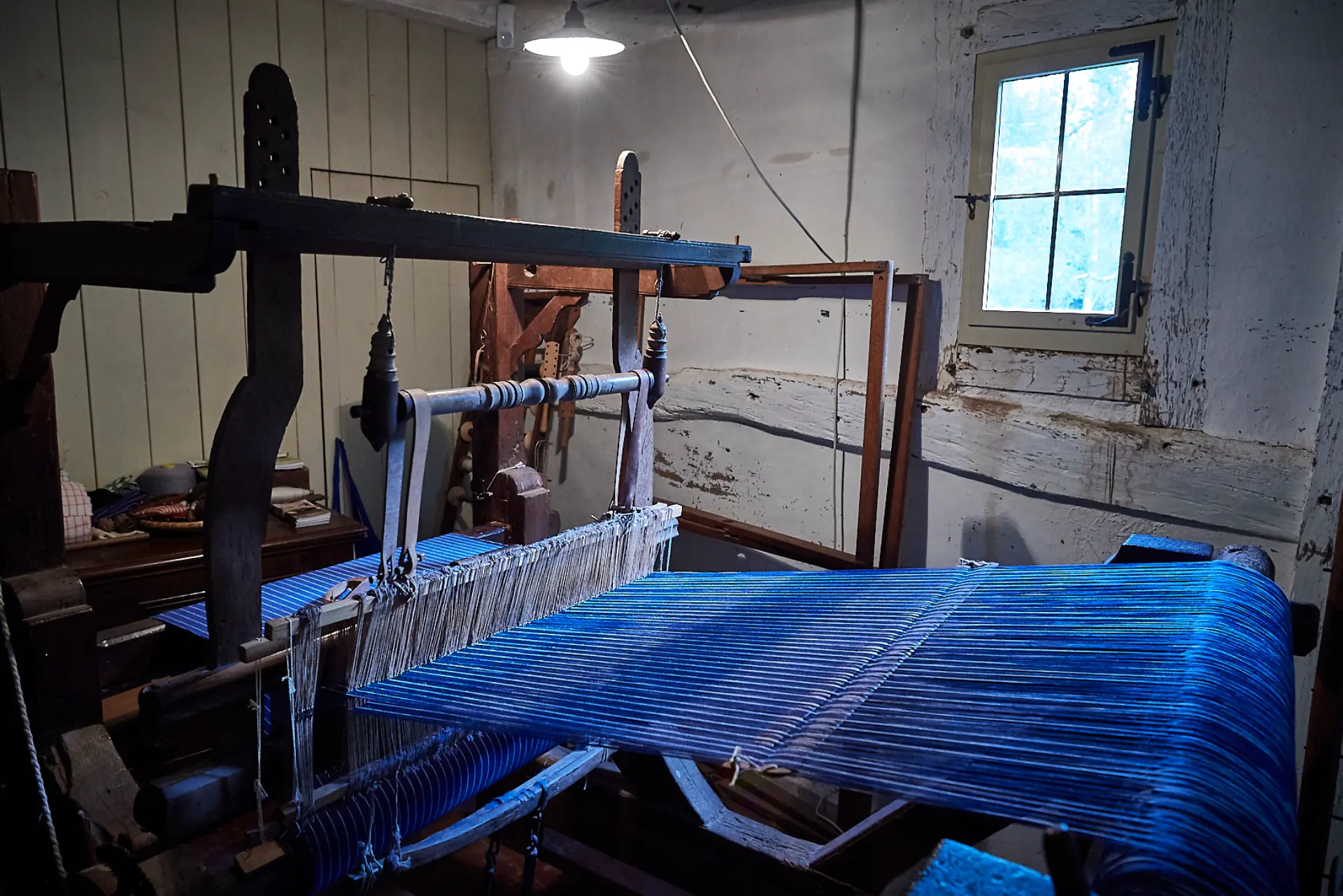 Eine ältere Frau sitzt an einem rustikalen Holztisch in einem historischen, steinernen Raum mit warmem Licht. Sie trägt runde Brille, ein graues Kopftuch, einen dunklen Schal und schlichte, traditionelle Kleidung. In ihren Händen hält sie einen drahtförmigen Gegenstand, der wie eine handgefertigte Mausefalle aussieht. Auf dem Tisch liegen weitere Drahtkonstruktionen sowie Werkzeuge wie Zangen.