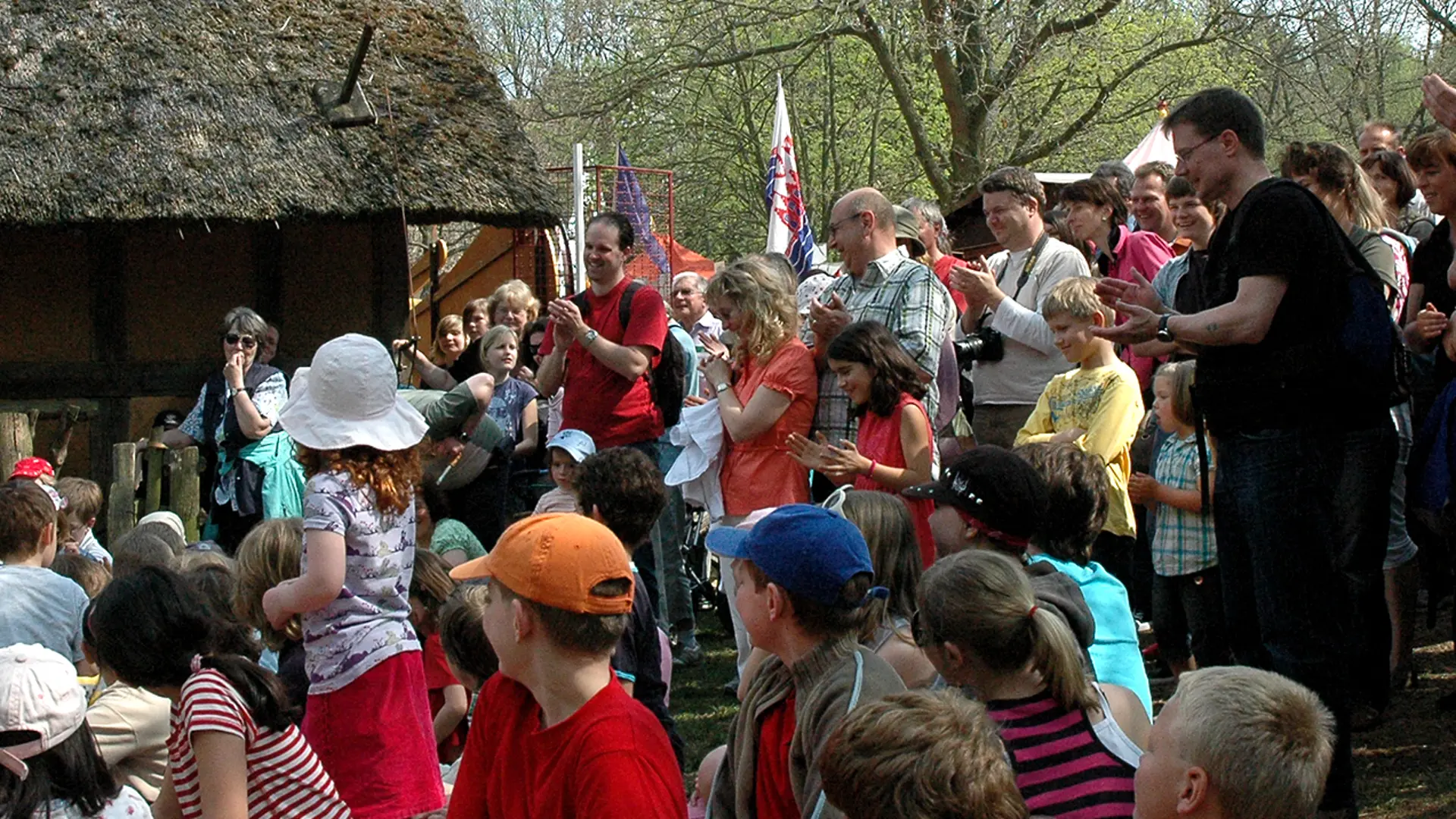 Foto: große Gruppe von Kindern und Erwachsenen im Freien vor einem strohgedeckten Gebäude.