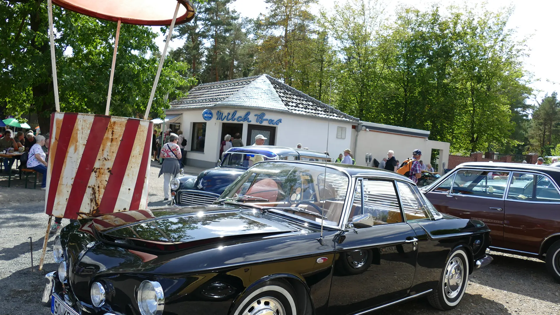 schwarzer Oldtimer mit glänzendem Lack, verchromten Stoßfängern und Zierleisten sowie Weißwandreifen. Das Fahrzeug steht auf einem befestigten Platz. Dahinter sind weitere historische Autos und ein weißes Gebäude mit der Aufschrift „Milch Bar“ zu sehen. Links steht ein rot-weiß gestreifter Verkehrslenkungsposten mit einem runden Dach.