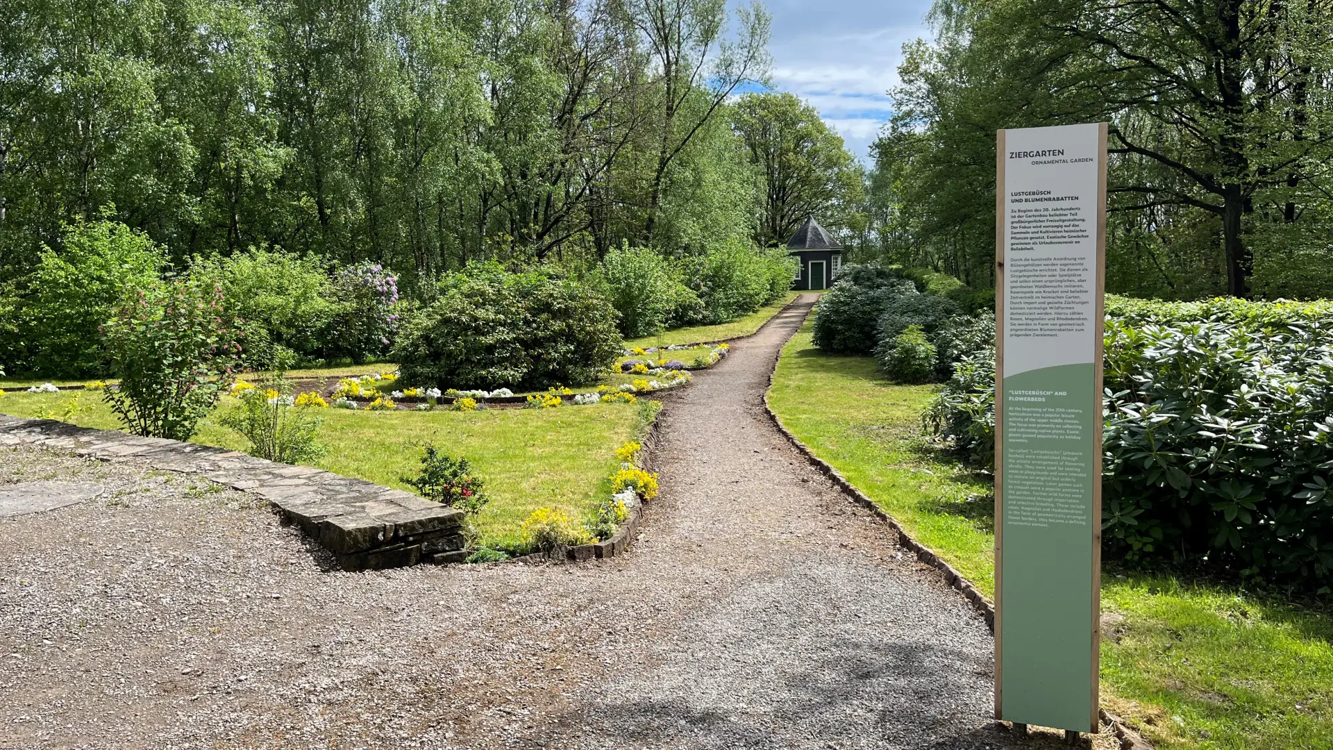 ein geschotterter Gartenweg führt durch einen grünen Garten mit Gras und hohen runden Büschen. Vorne rechts im Bild steht eine Säule mit Erklärtext zum Garten.